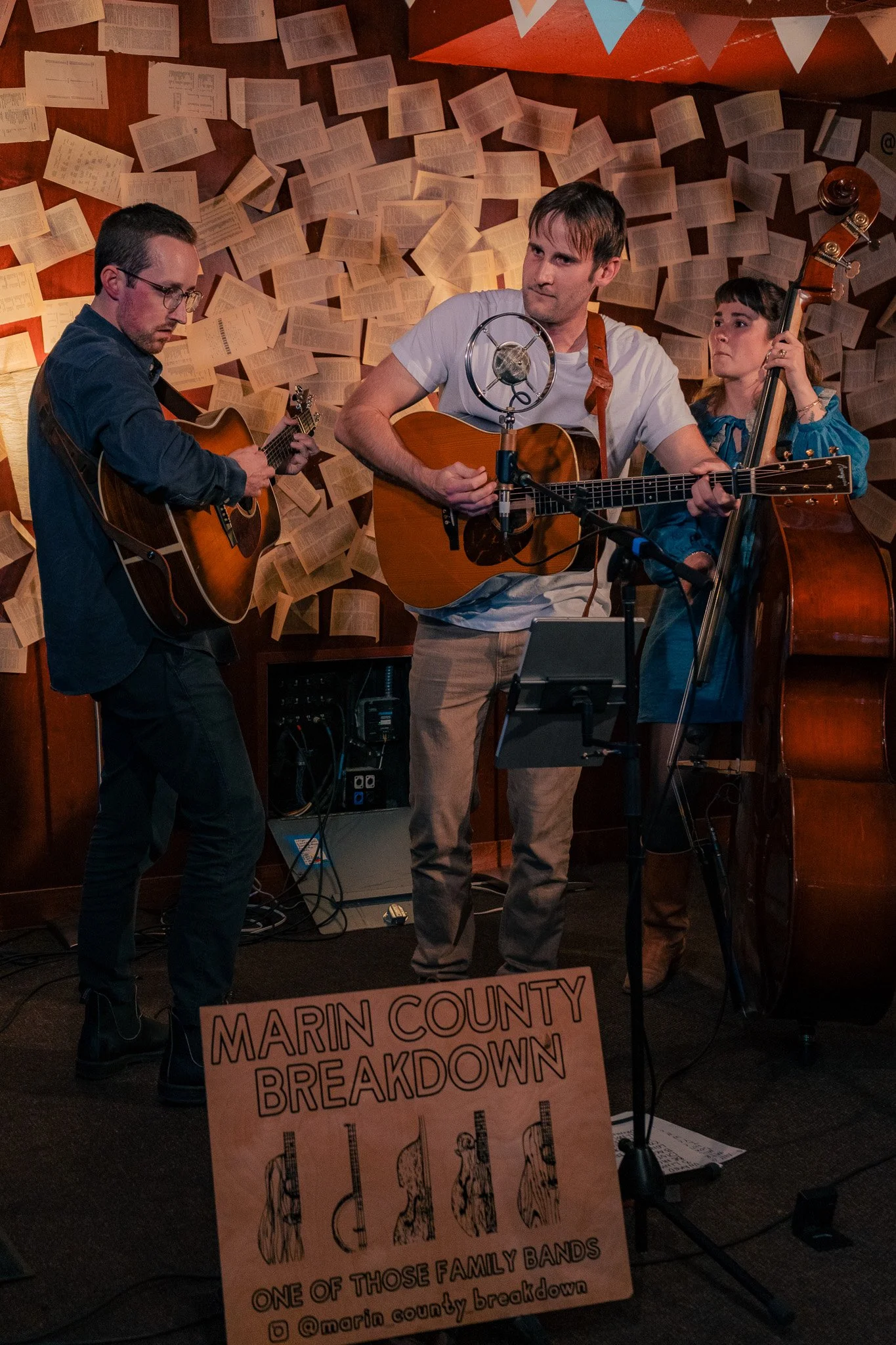 Three musicians performing on stage with guitars and a double bass, in front of a wall decorated with pages from books and a sign that reads 'Marin County Breakdown' with drawings of guitars and the phrase 'One of those family bands'.
