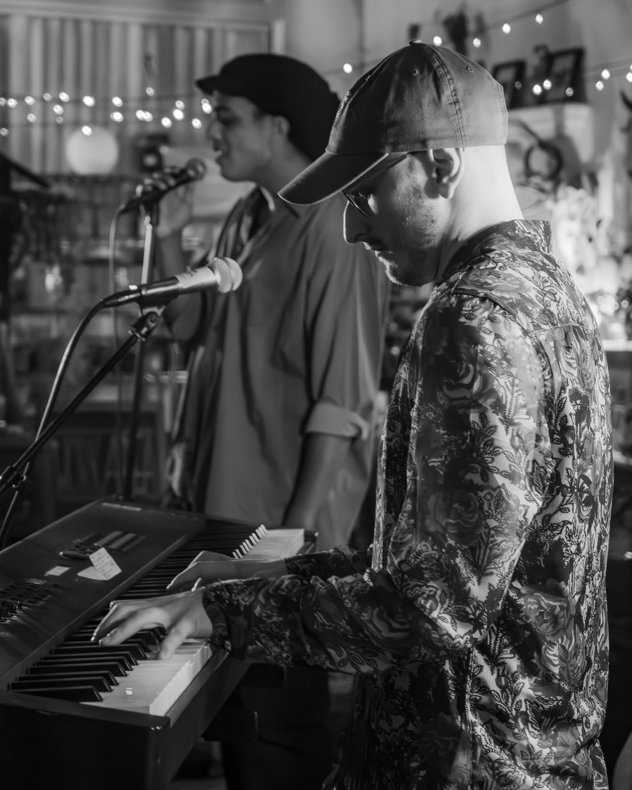 Two musicians performing indoors, one playing keyboard and the other singing into a microphone, with decorative string lights in the background.
