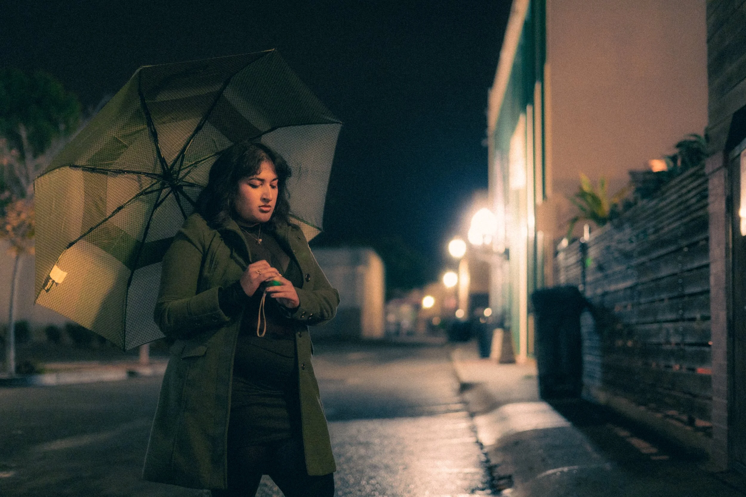 A woman in a green coat holding an umbrella and looking at her cell phone on a rainy night on a city street.