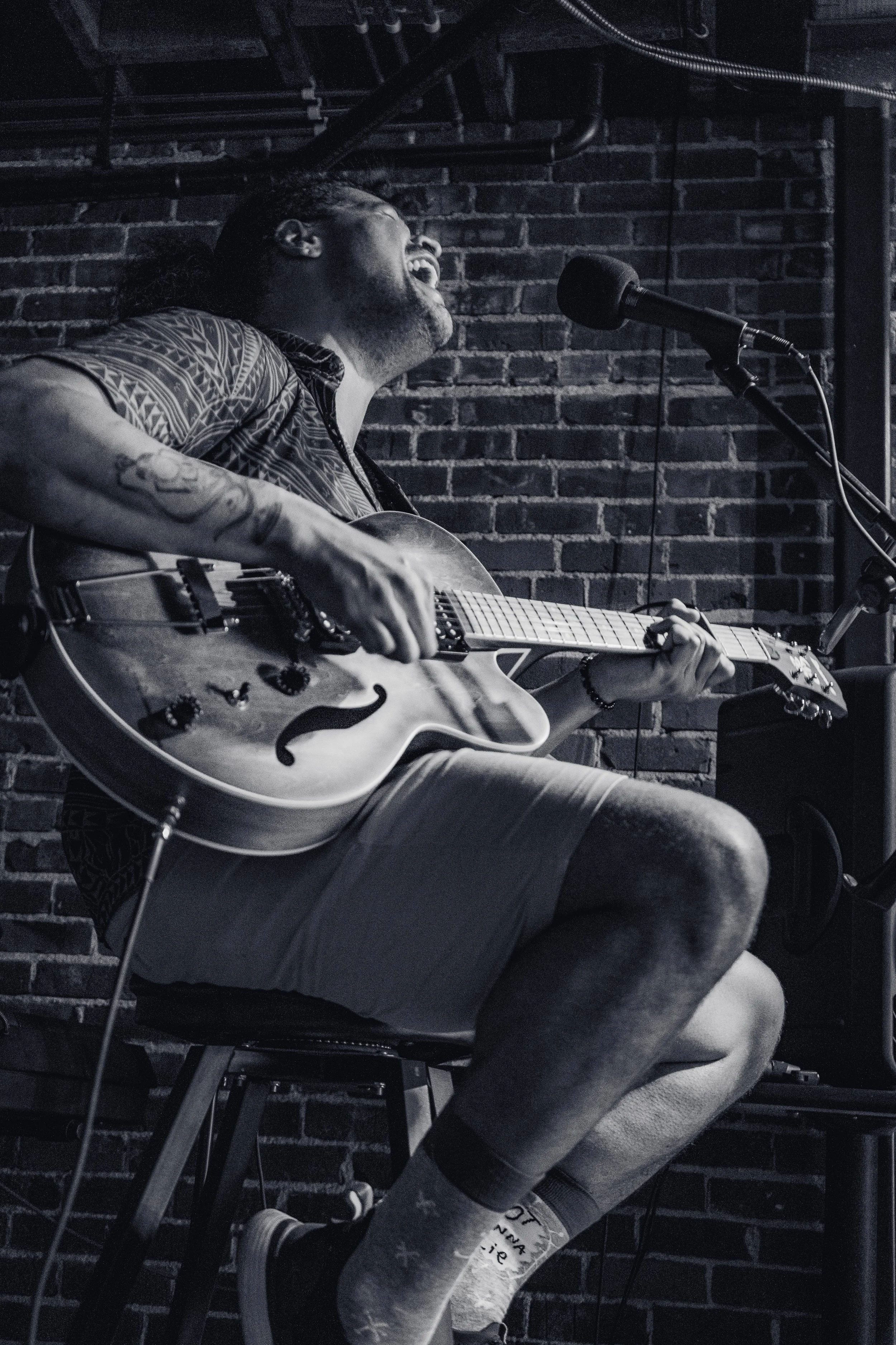 A man singing passionately while playing an acoustic guitar on stage in front of a brick wall.