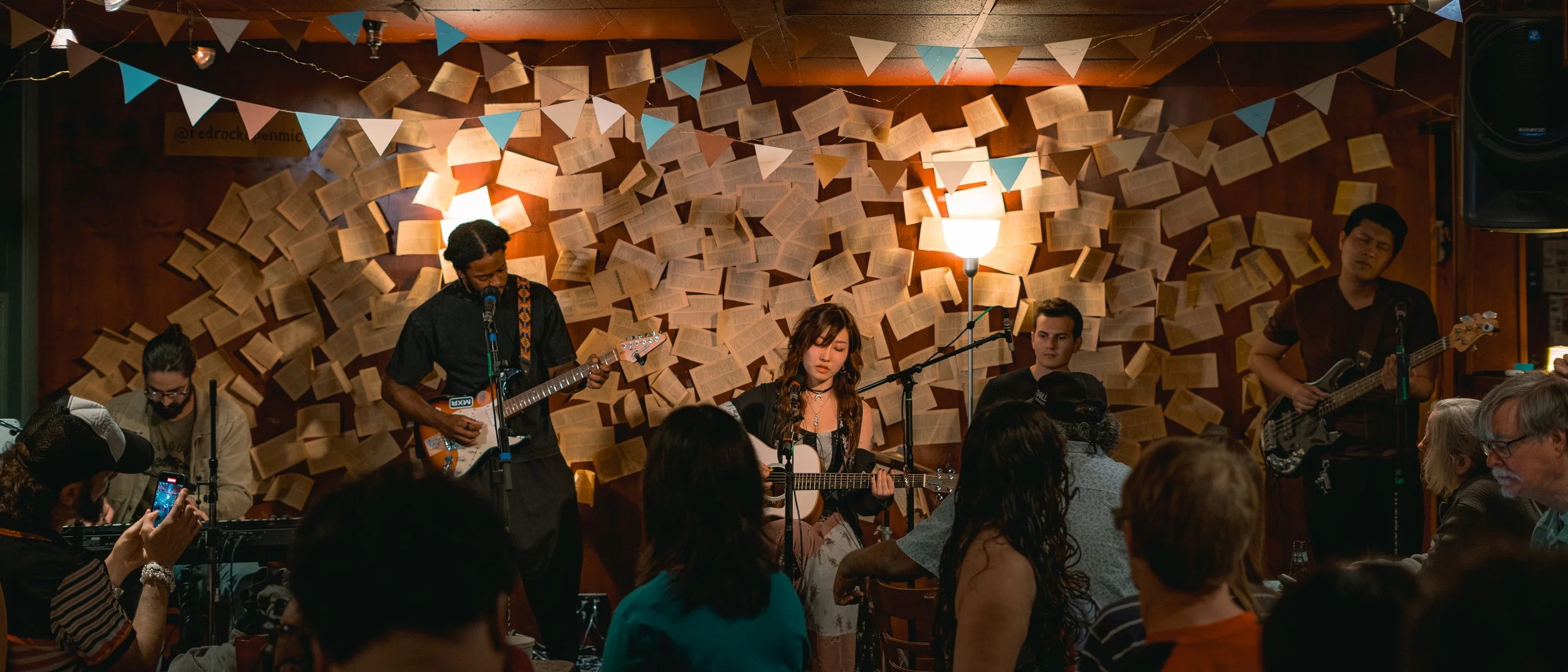 Band performing live at a cozy venue with open books on the wall and colorful bunting overhead