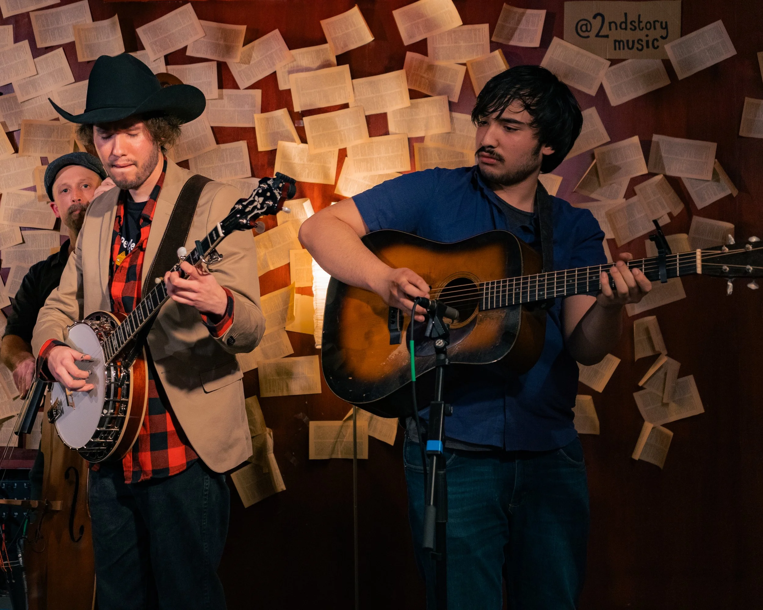 Two men playing guitars, one with a banjo and the other with an acoustic guitar, in front of a backdrop of open books pinned to the wall. The scene appears to be a live music performance in a cozy, literary-themed venue.