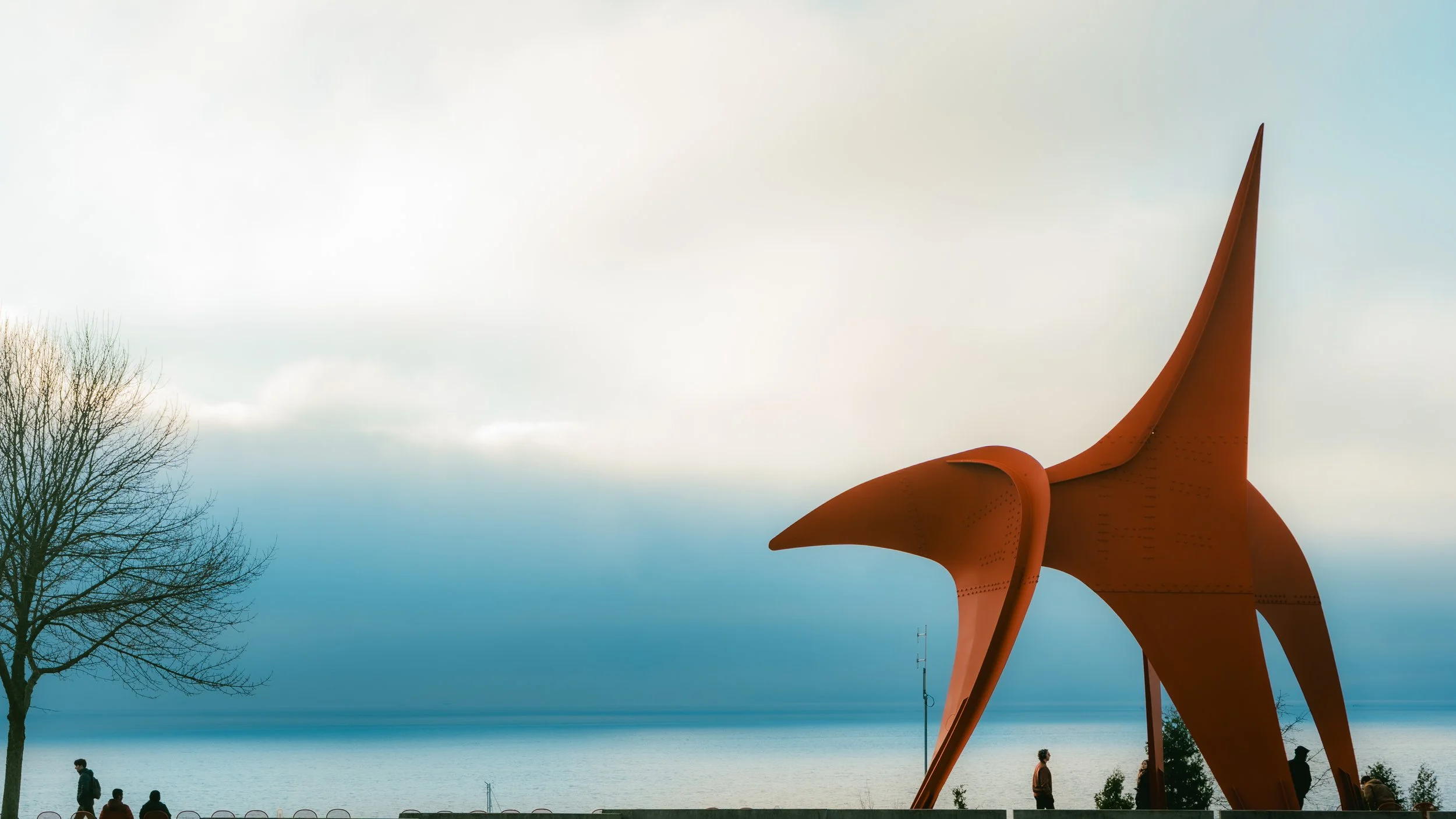 Large abstract sculpture resembling a bird in flight, located near a body of water on a cloudy day with a few people walking nearby.