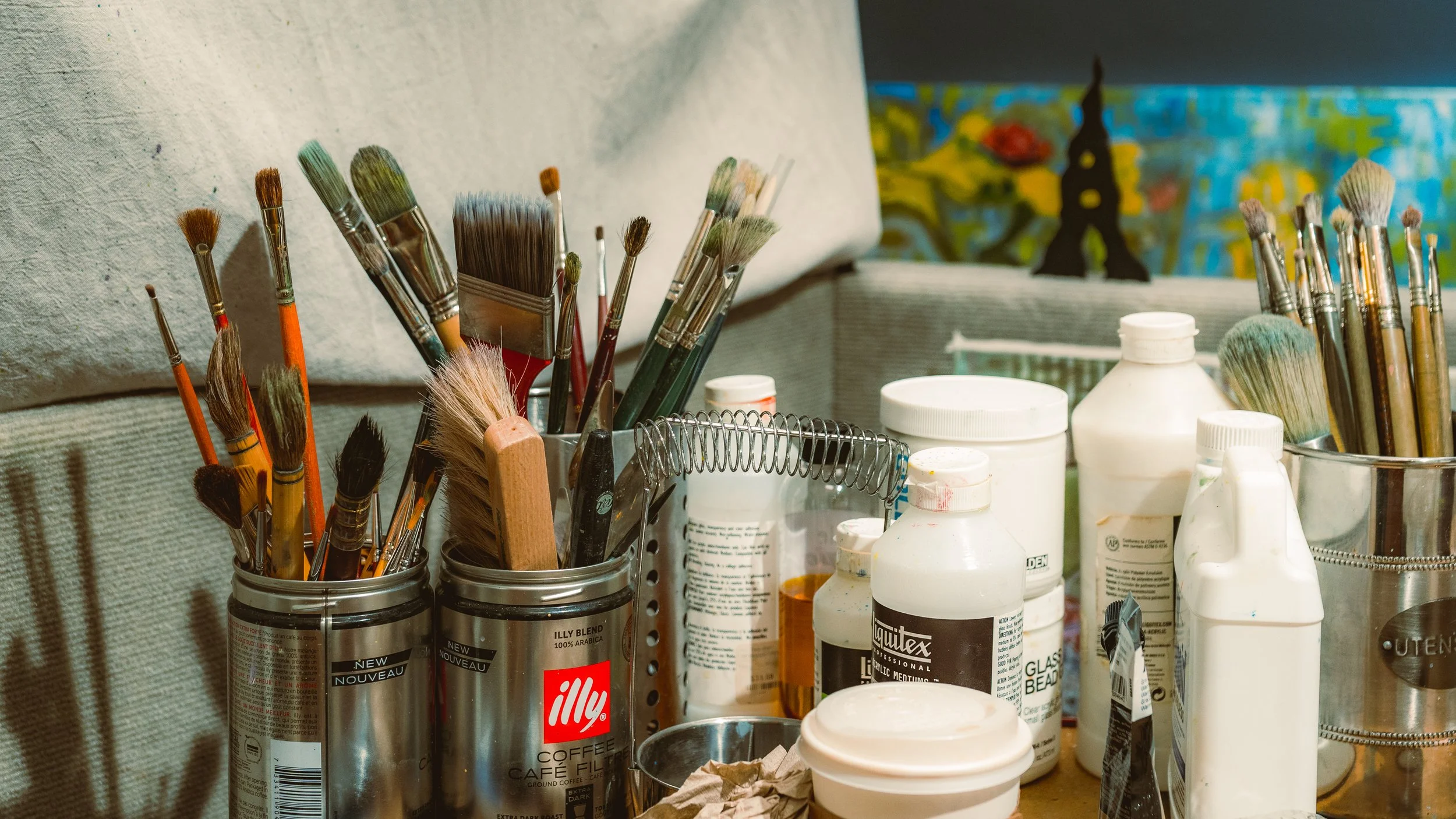 A collection of paintbrushes and painting supplies on a table, with a couch and colorful artwork in the background.