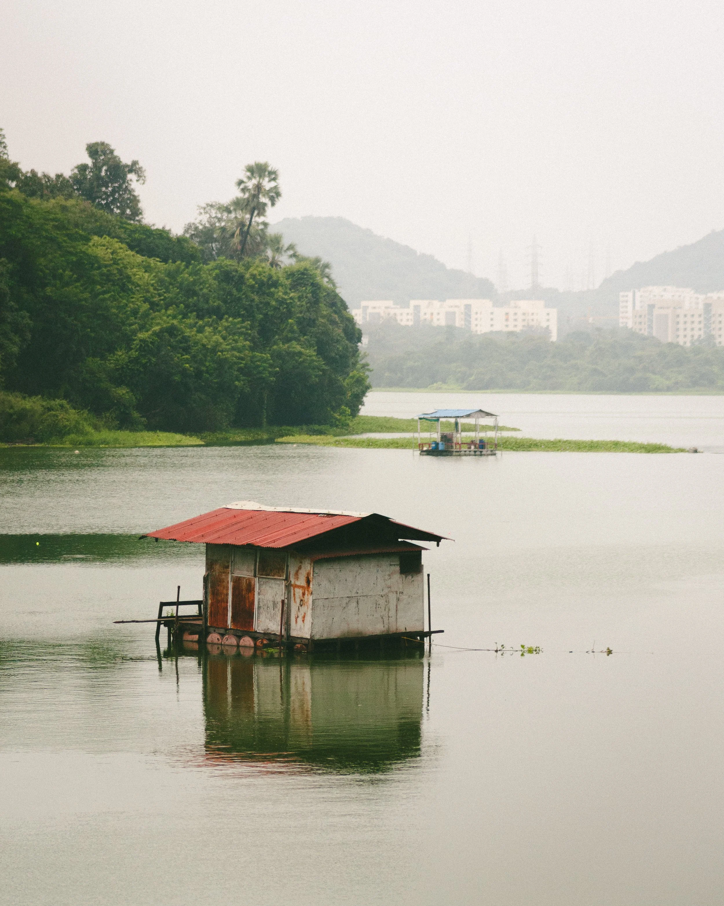 A floating house with a red roof on a calm lake, with a lush green hillside and distant buildings in the background.