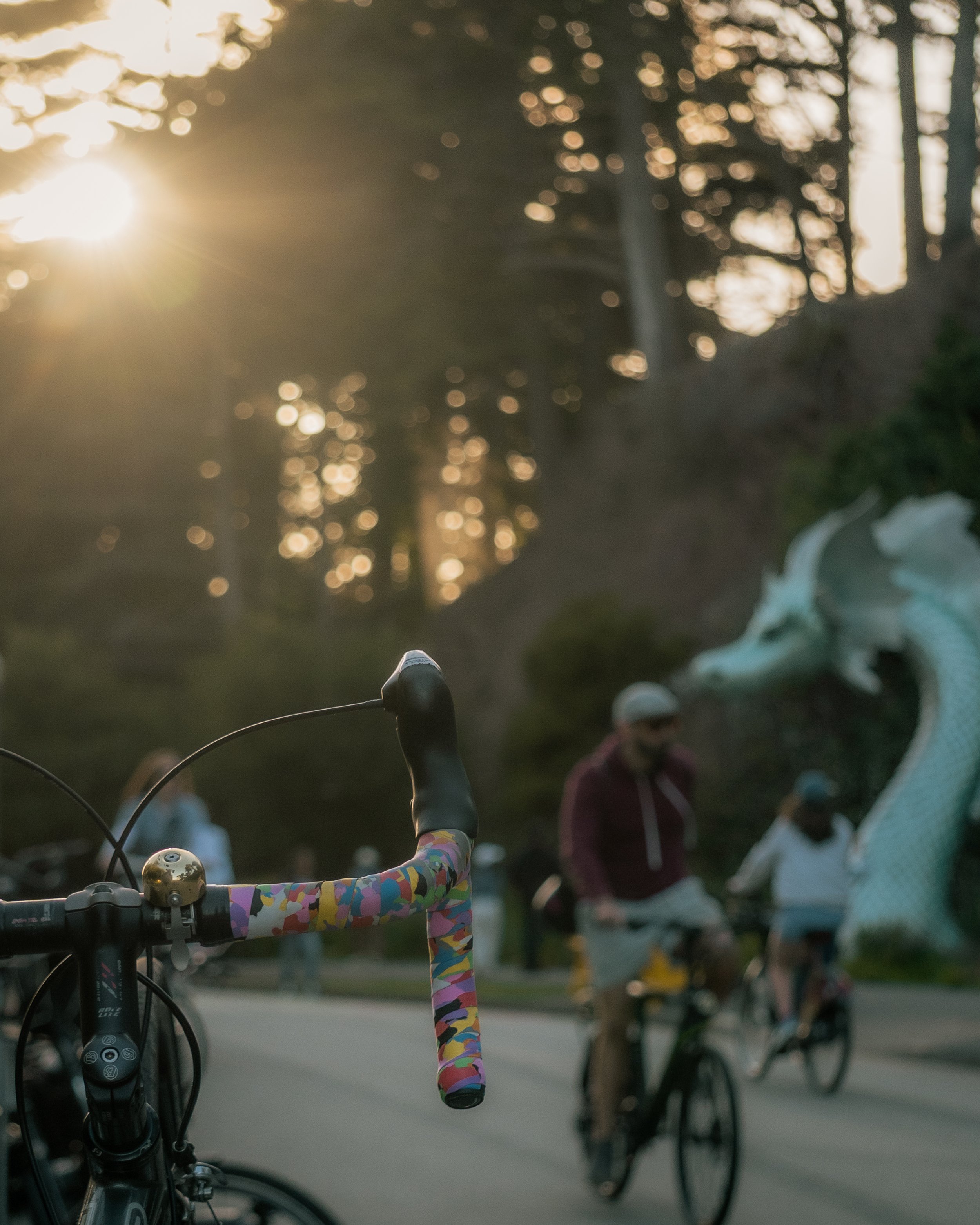A close-up view of a bicycle handlebar with a colorful, patterned tape and a bell, with two people riding bicycles in the background near a dragon sculpture, under a setting sun in a wooded area.