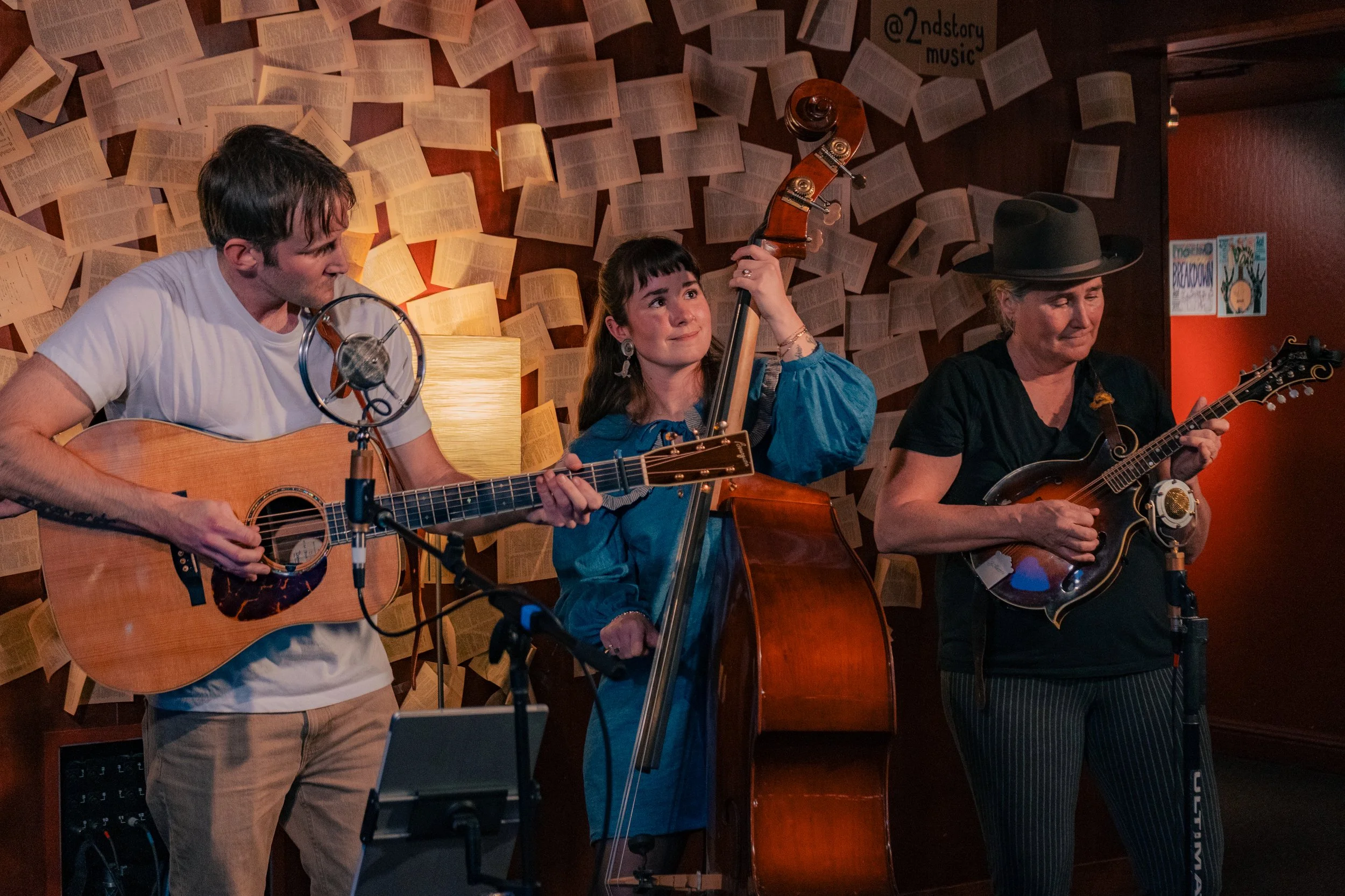 Three musicians performing in a dimly lit room with a background of open books on the wall. The man on the left is playing an acoustic guitar and wearing a white t-shirt. The woman in the middle is playing a double bass and wearing a blue dress with 