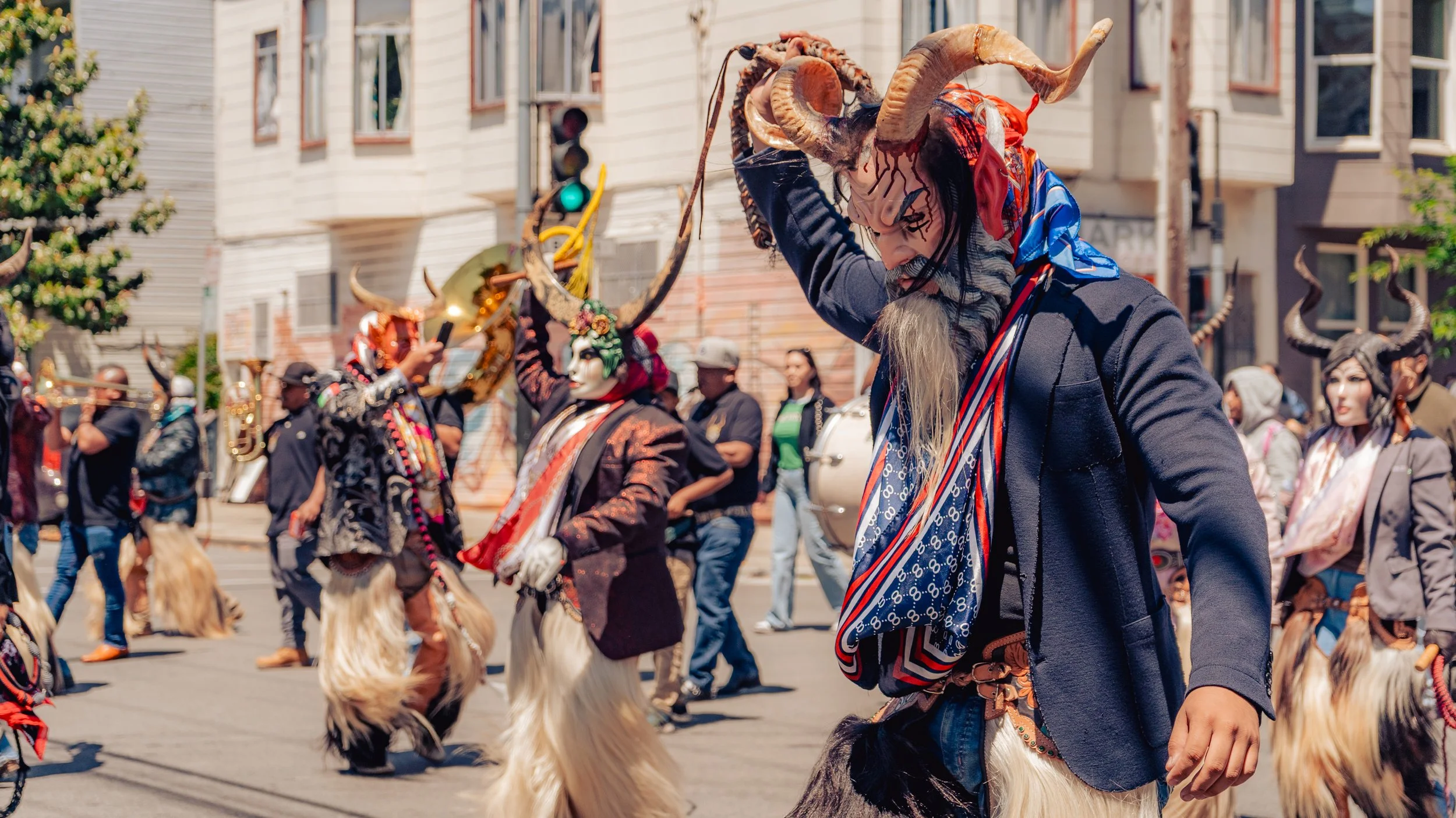 People in costumes participating in a parade, wearing horned masks and animal-like clothing, dancing and playing instruments in an urban street.