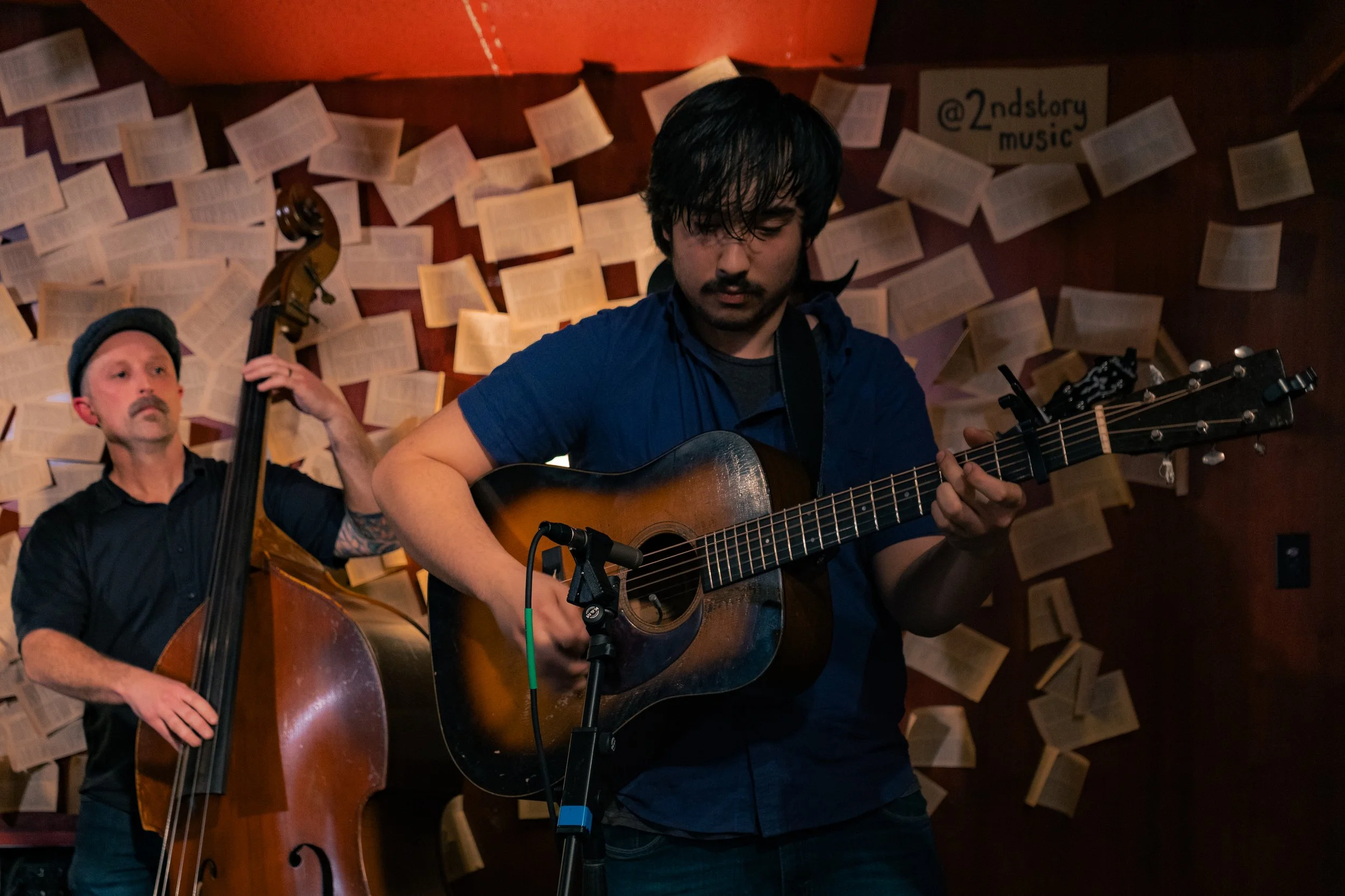 Two musicians performing with guitars in a dimly lit room decorated with pages of open books pinned to the wall.