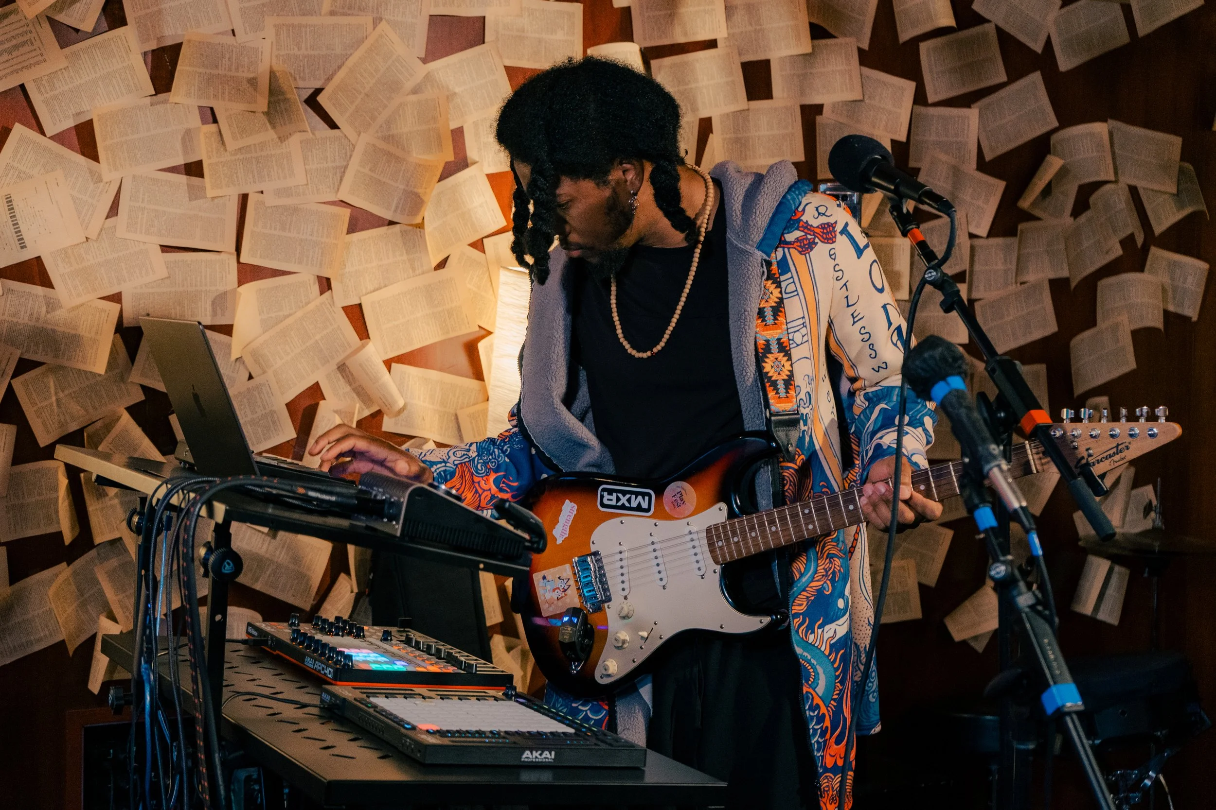 Musician with dreadlocks playing electric guitar, surrounded by electronic music equipment, in a room decorated with open books on the wall.
