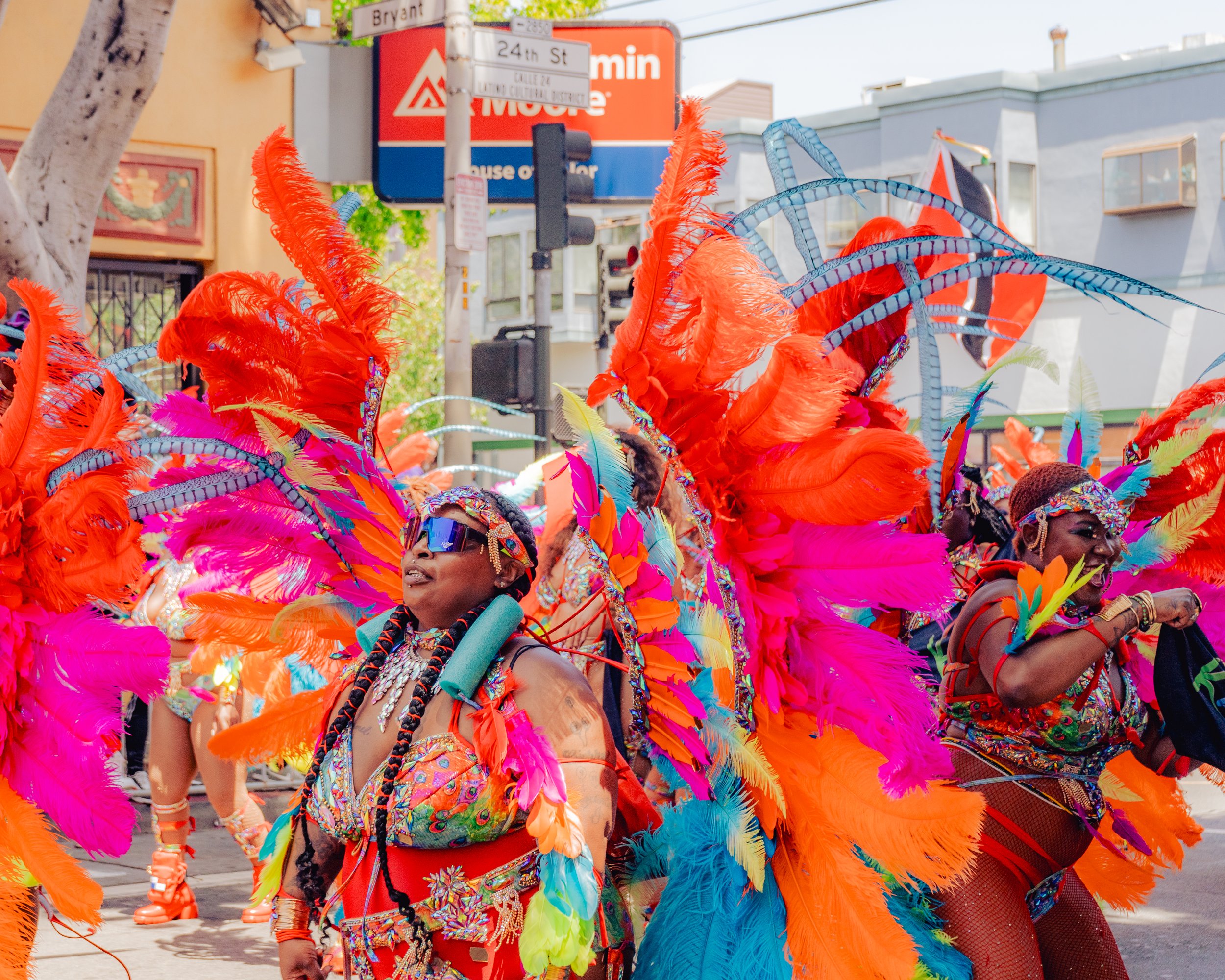 Group of people in colorful feathered costumes participating in a parade, with storefronts and street signs in the background.