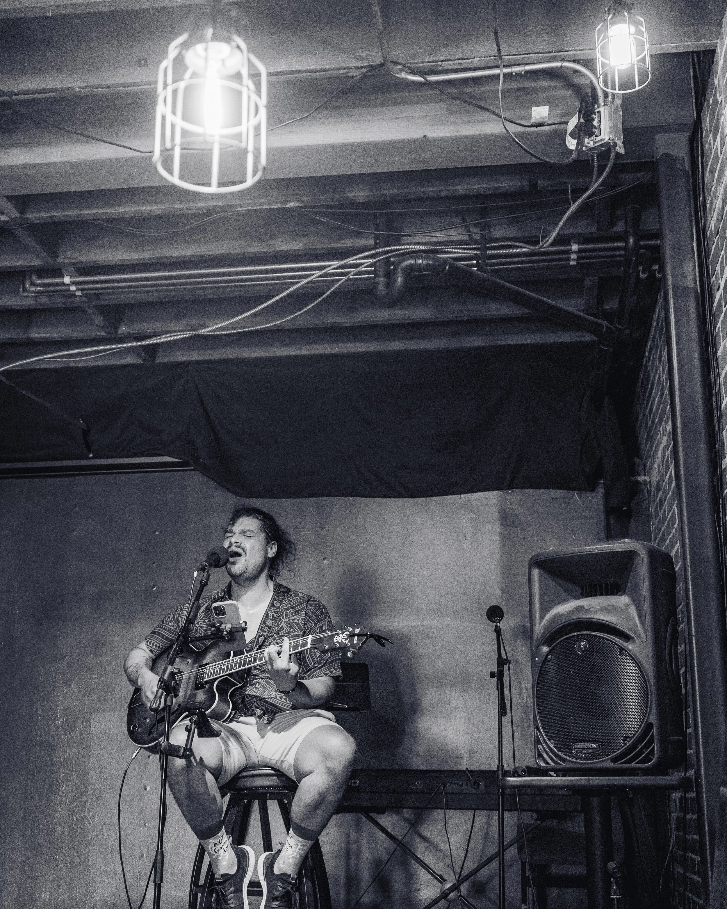 A man singing and playing an acoustic guitar on a small stage, with a microphone in front of him. The background features a plain wall and a large speaker on his right. The scene appears to be in a dimly lit, intimate venue with industrial decor.