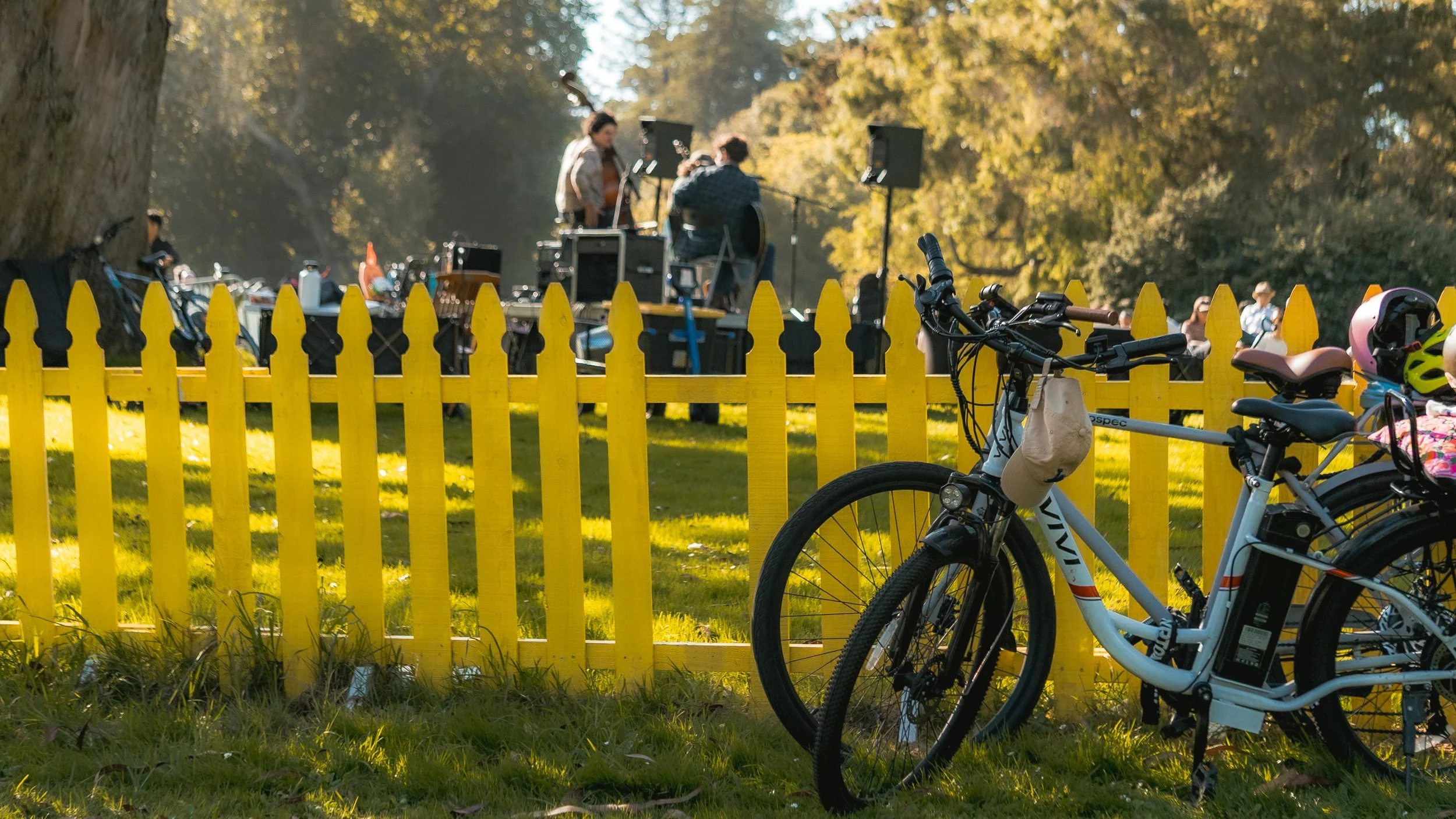 Bicycles parked by a bright yellow picket fence with people gathered around a small outdoor stage or setup in the background during sunset.