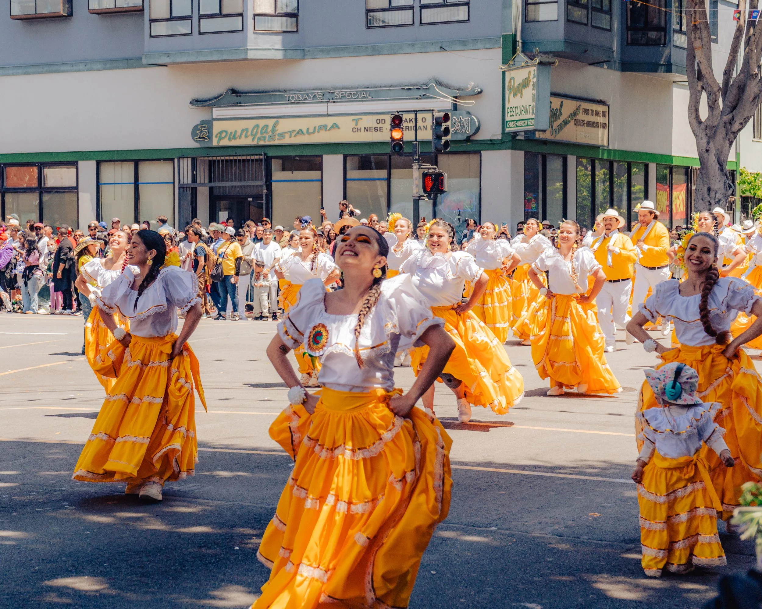 A parade with women and children in bright yellow skirts and white blouses performing a dance in the street. Spectators watch from the sidewalk, behind a traffic light, in front of a building with signs for restaurants.