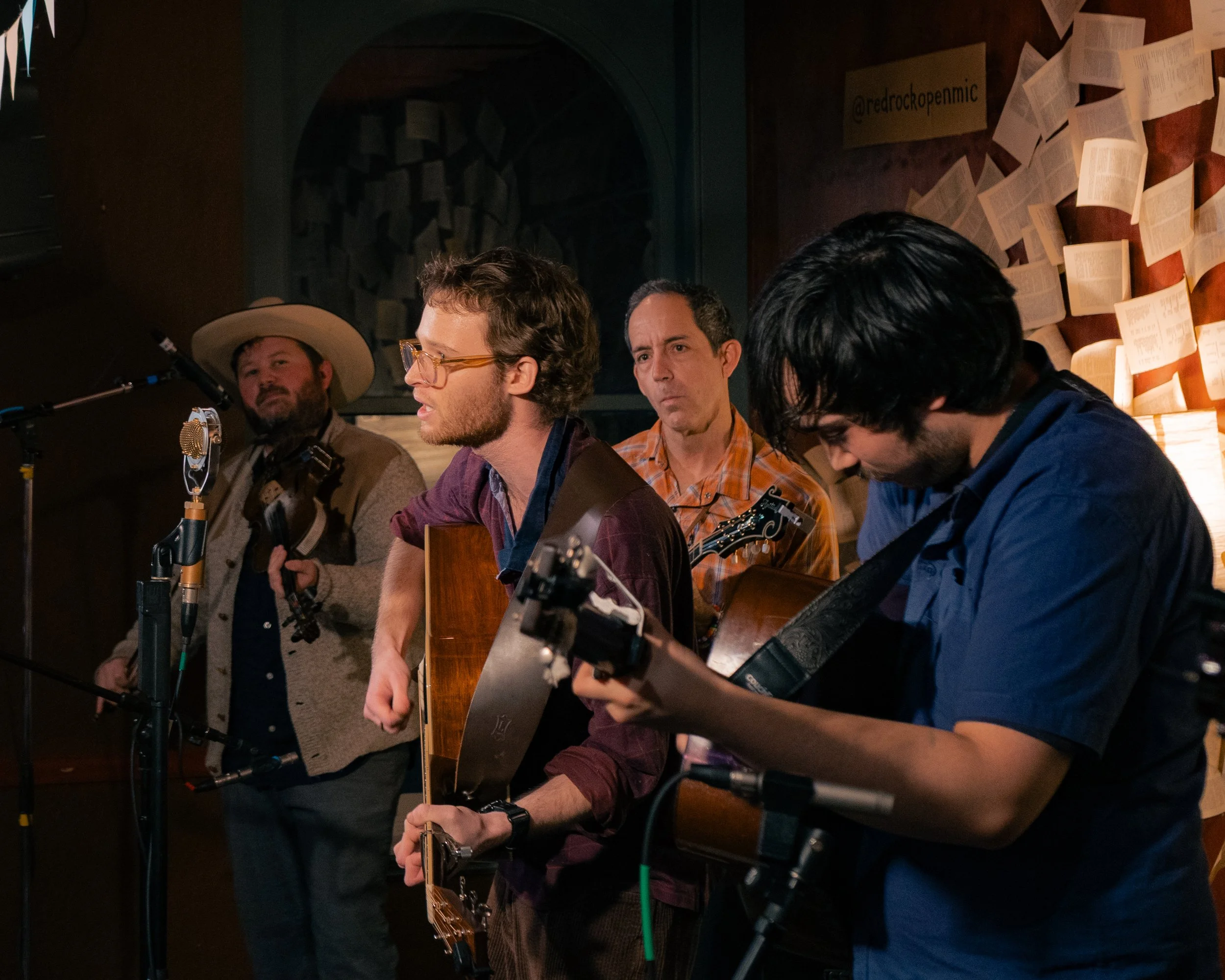 Four men are performing music in a dimly lit room, with one singing into a microphone, two playing guitars, and the fourth holding a guitar strap.