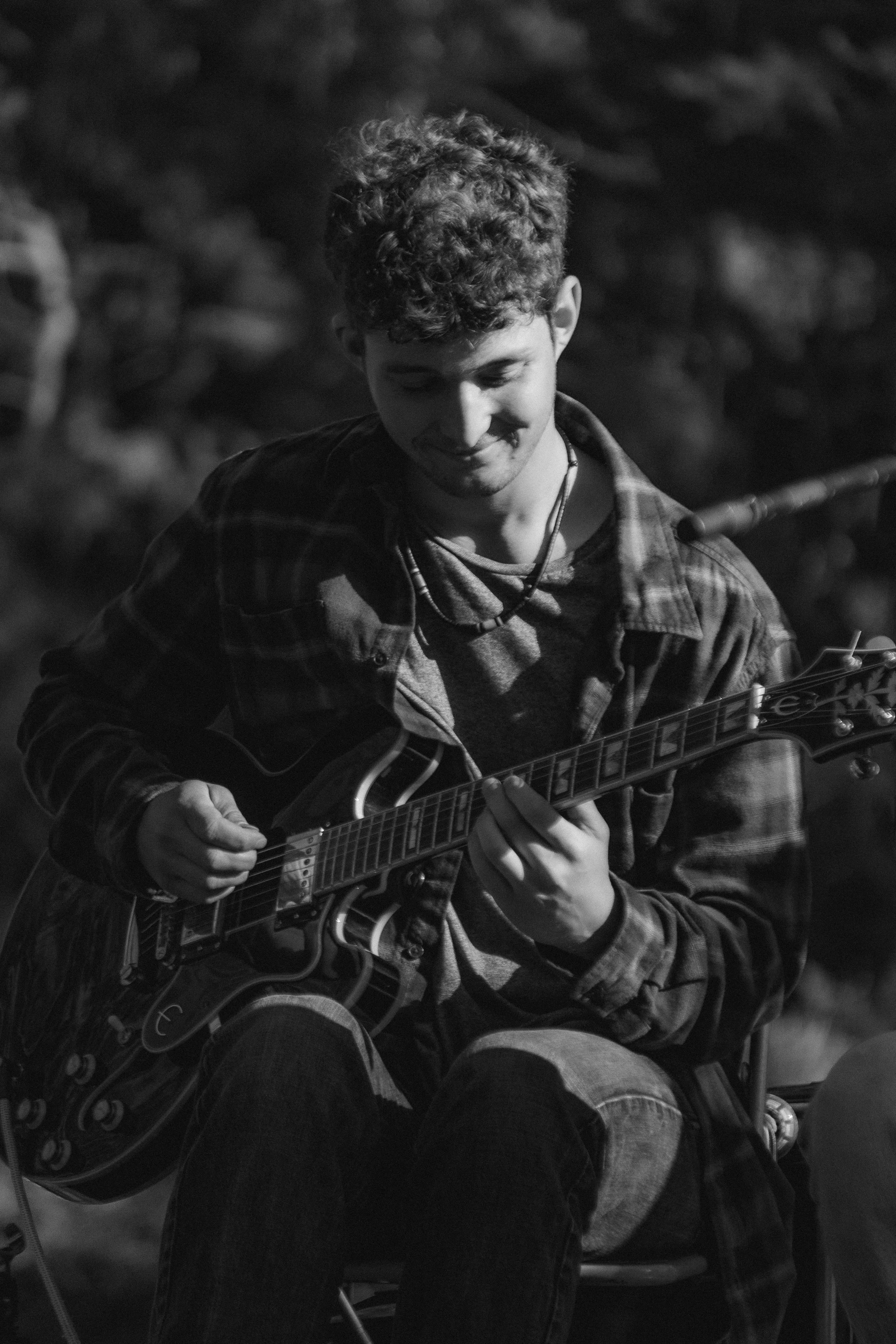 A young man with curly hair playing an electric guitar, wearing a plaid shirt and sitting outdoors at night.