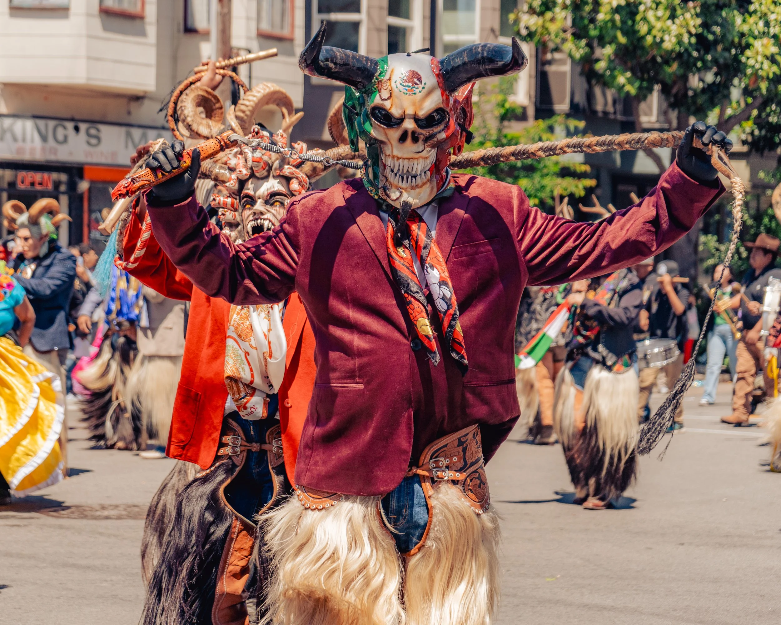 A person dressed in a festive costume with a skull mask and horns, carrying a staff, participating in a parade with others dressed in colorful costumes and masks.