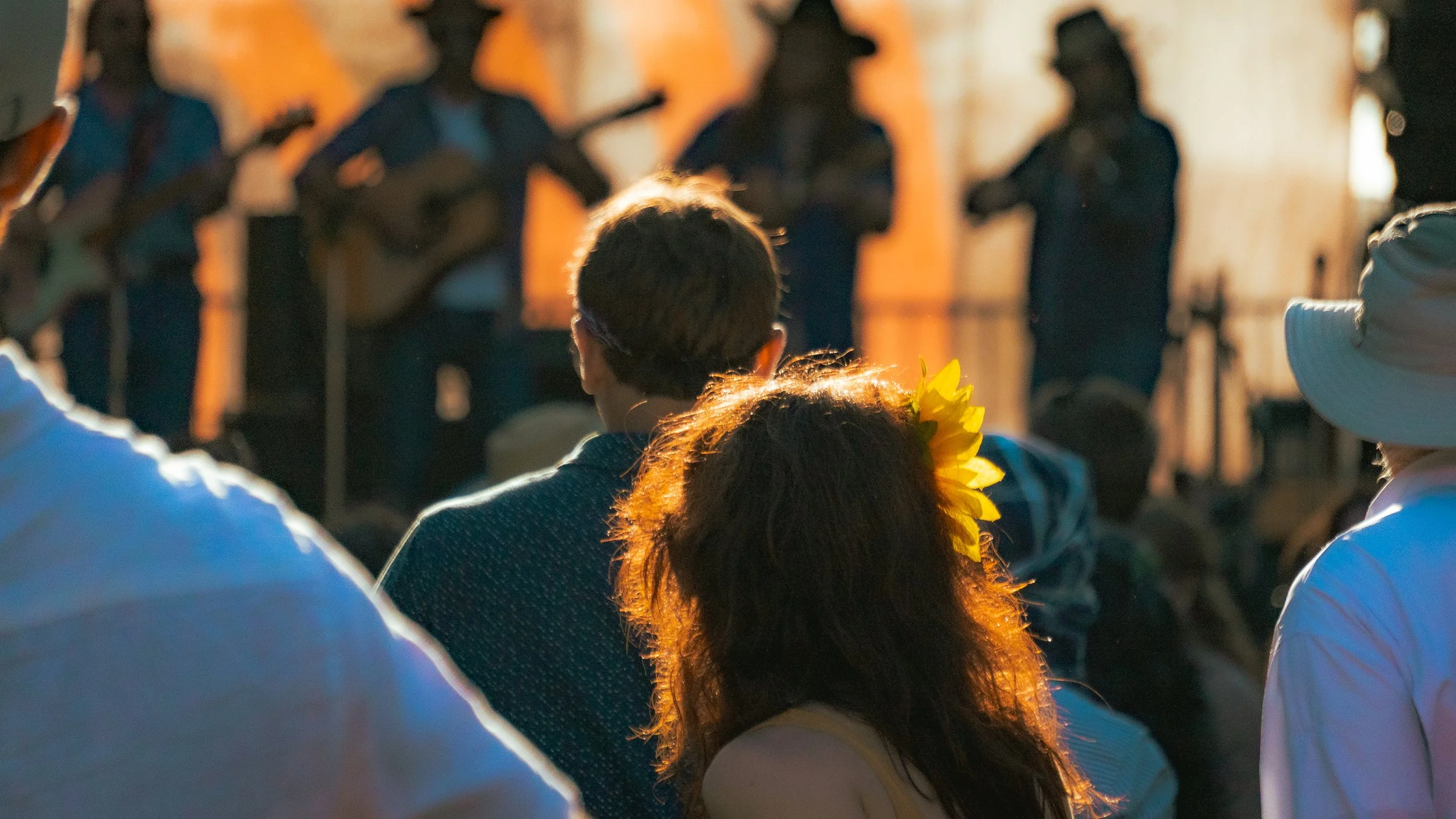 Back view of a person with curly reddish hair and a yellow flower in her hair, watching a musical performance on stage. Other audience members are visible, some wearing hats, as musicians play guitars and sing in the background.