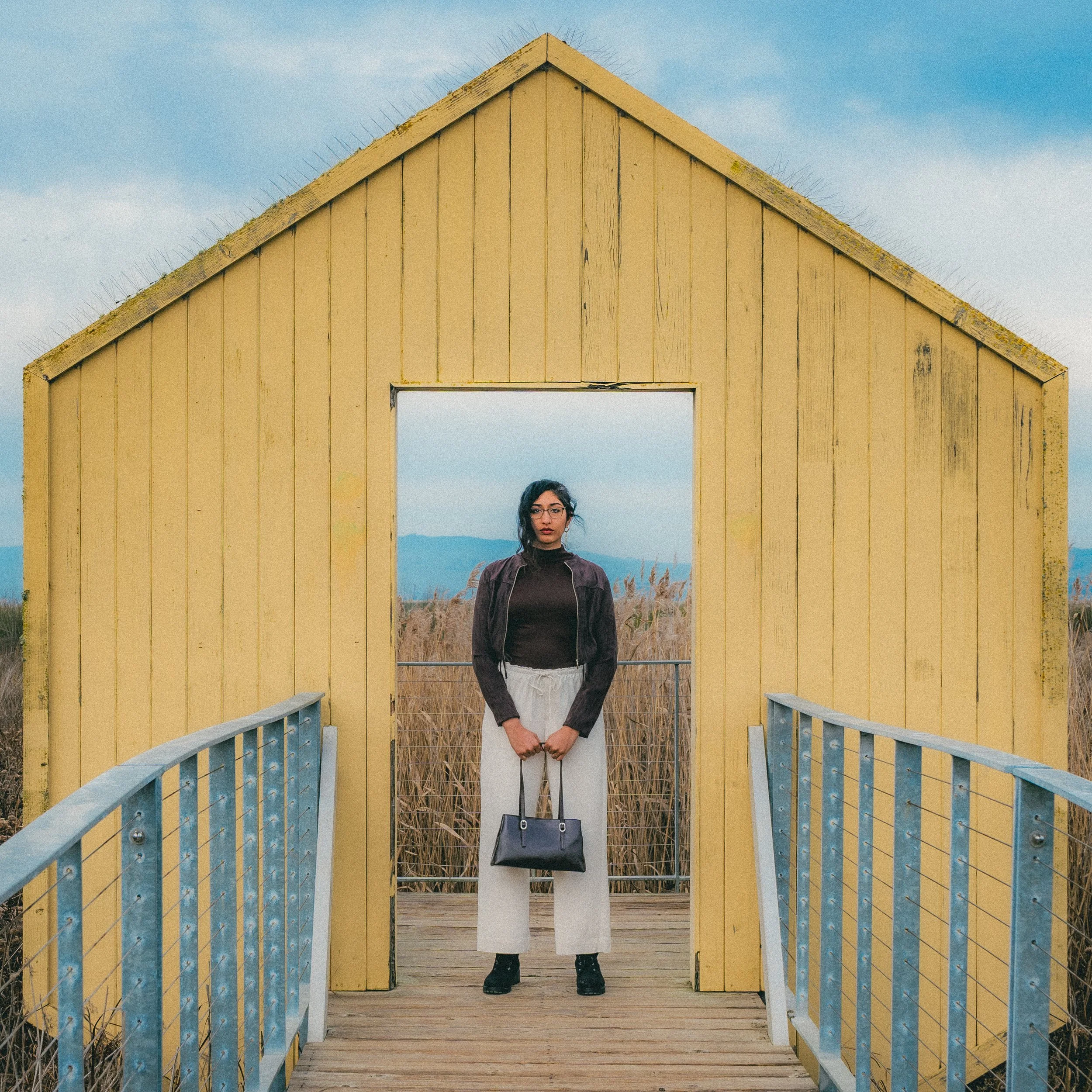 A woman with glasses and dark hair stands on a wooden bridge in front of a yellow wooden structure with a roof, holding a purse. The scene is outdoors with dry grass and mountains in the background under a blue sky.