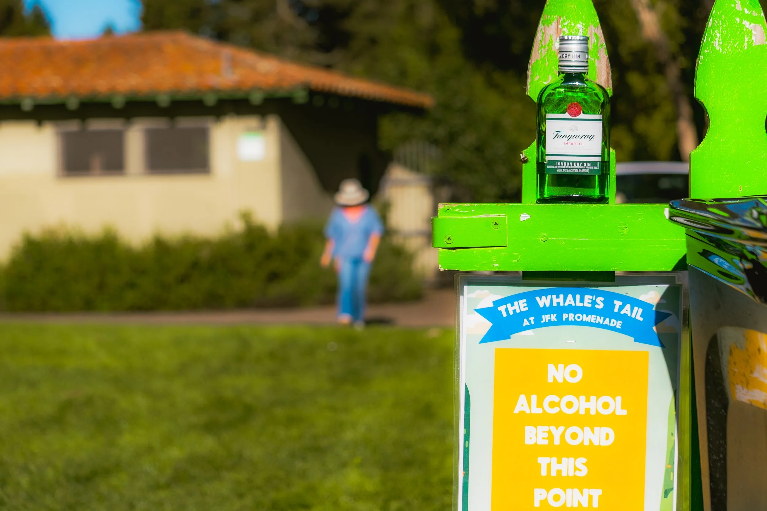 A sign at JFK Promenade with the phrase "No alcohol beyond this point" and a bottle of Tanqueray London dry gin on a bright green stand in the foreground. In the background, there is a person wearing a hat and blue clothing walking near a small build