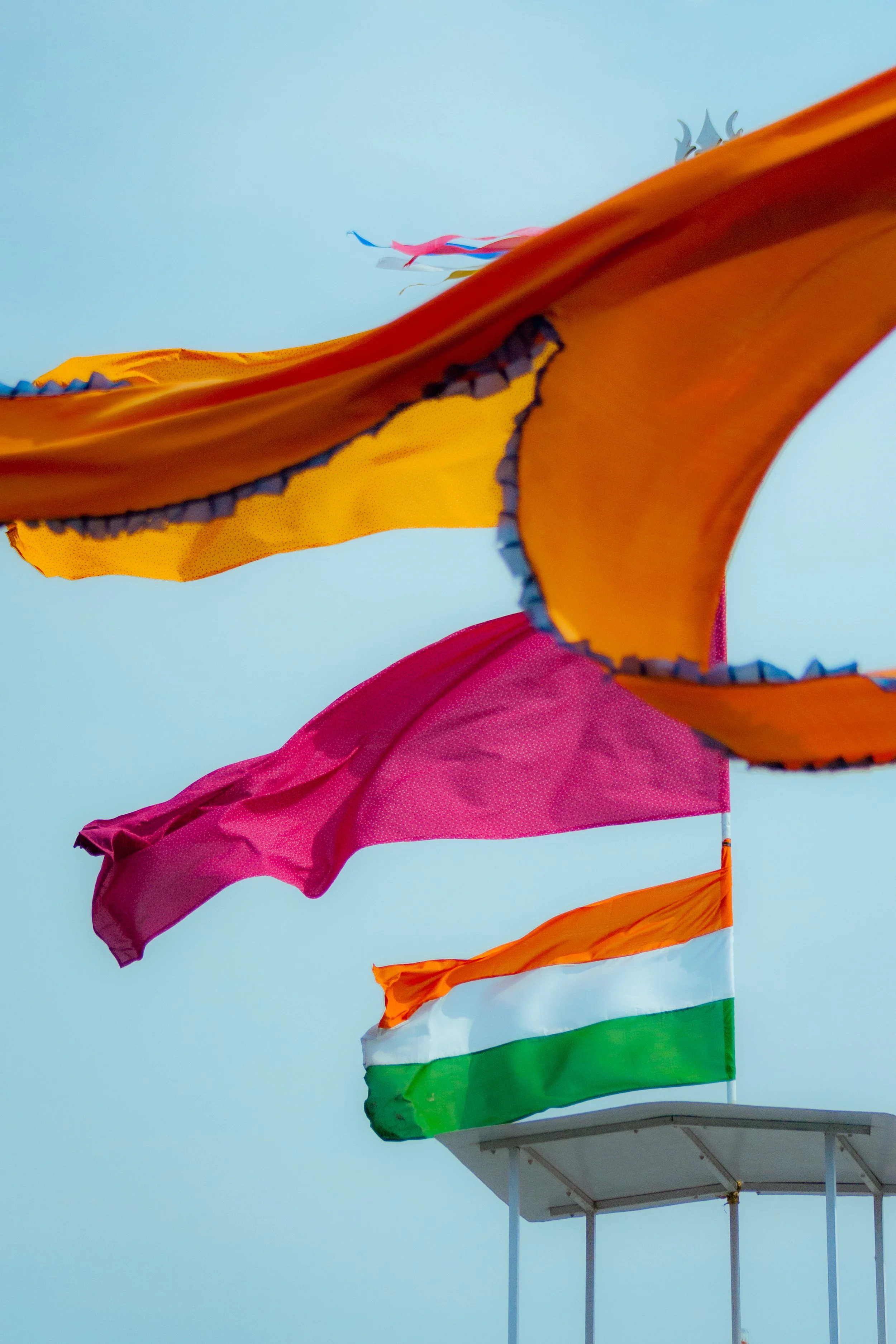 Multiple colorful flags, including the Indian flag, waving in the wind against a cloudy sky.