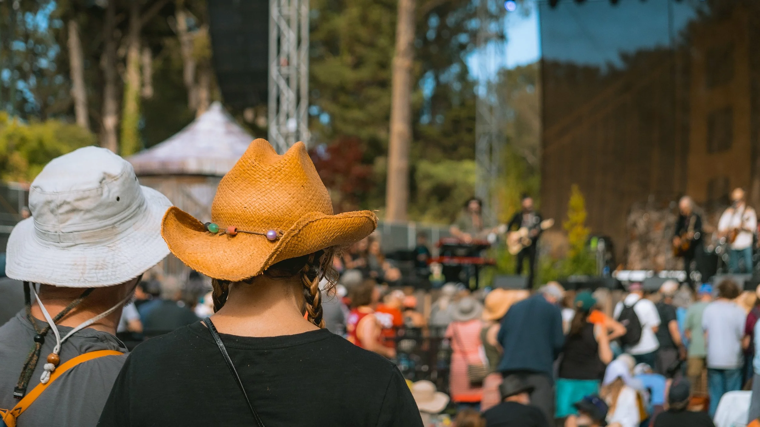 People attending an outdoor music festival, with two women in the foreground wearing hats, watching a band perform on stage, surrounded by trees and a crowd of festival-goers.