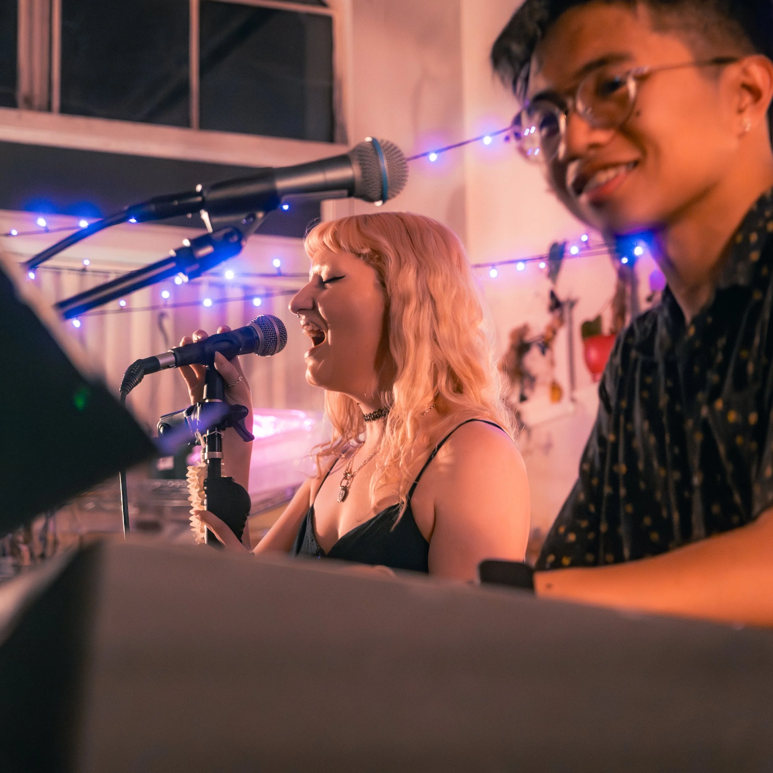 Two singers, a woman with blonde hair and a man with glasses, singing into microphones during a performance, with colorful string lights in the background.