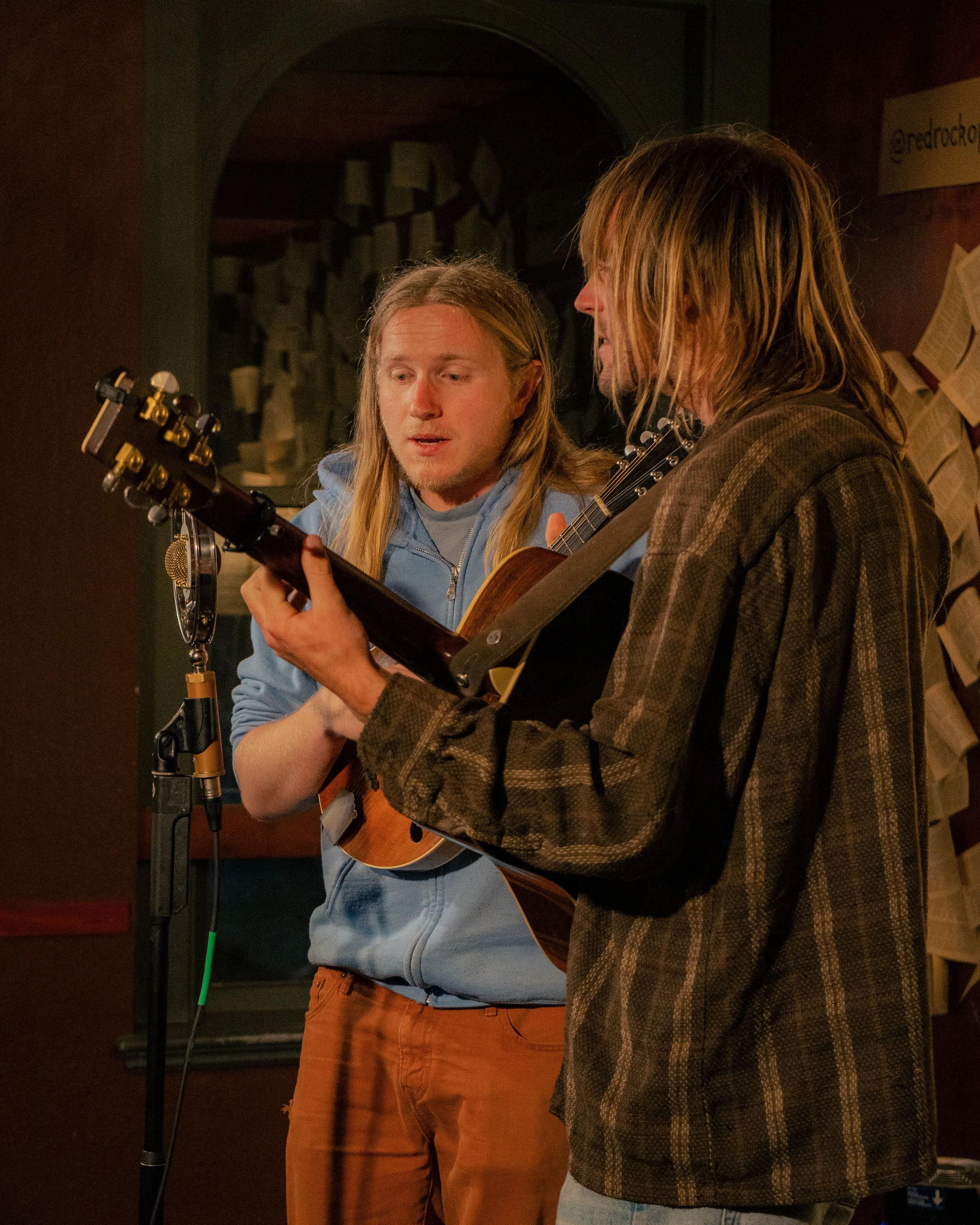 Two men with long hair playing guitars together in a recording studio or music room.
