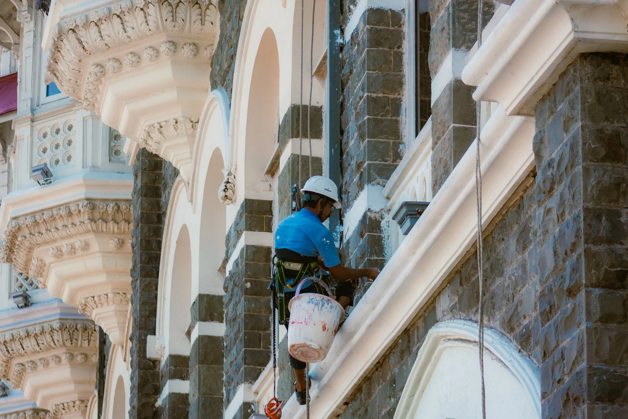 A worker in a blue shirt, white helmet, and safety harness cleaning the exterior of a historic building with ornate architectural details, using a bucket and cleaning tools, while suspended on ropes.