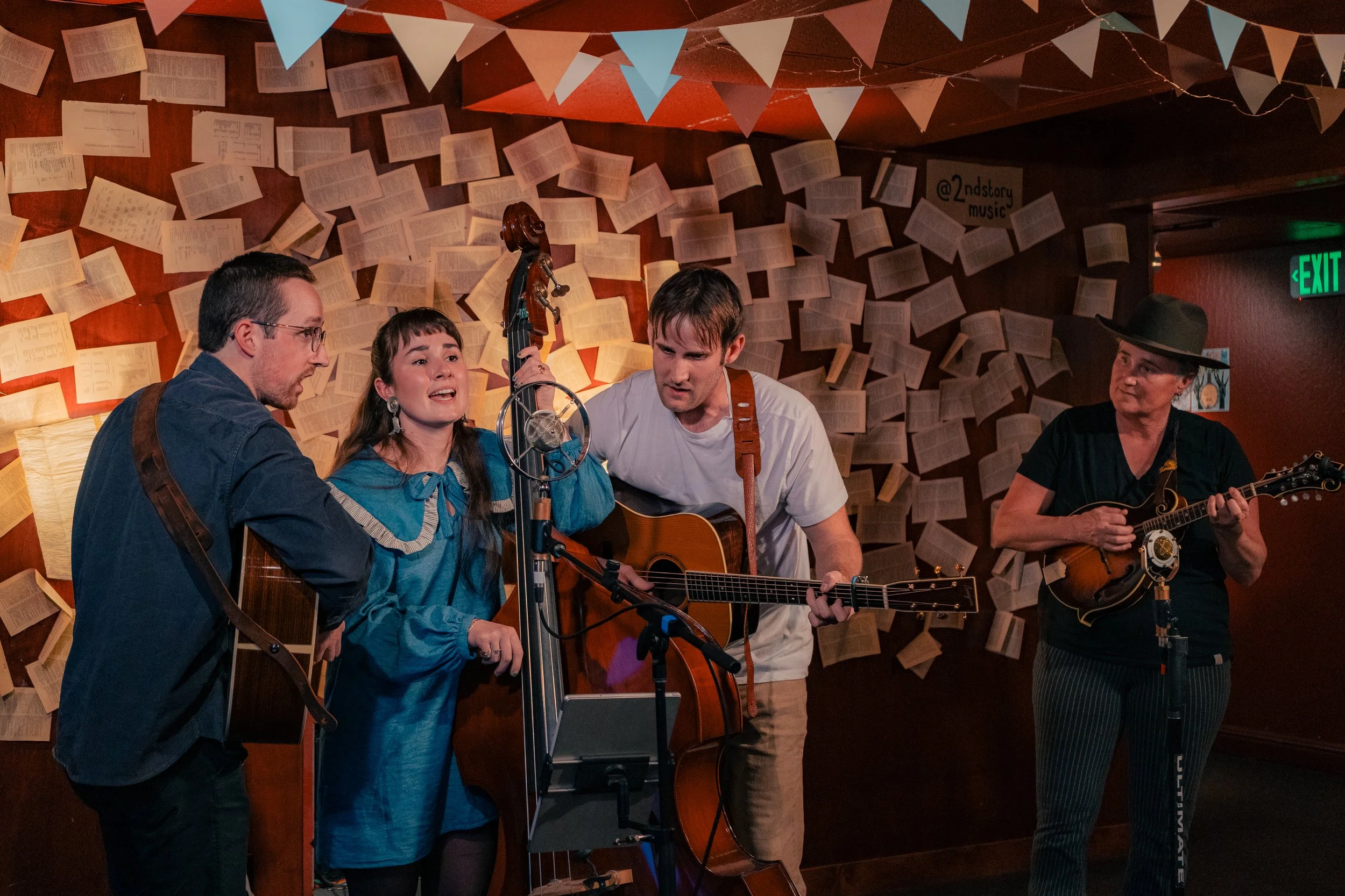 Four musicians performing with guitars and a mandolin in a decorated room with papers on the wall and bunting.