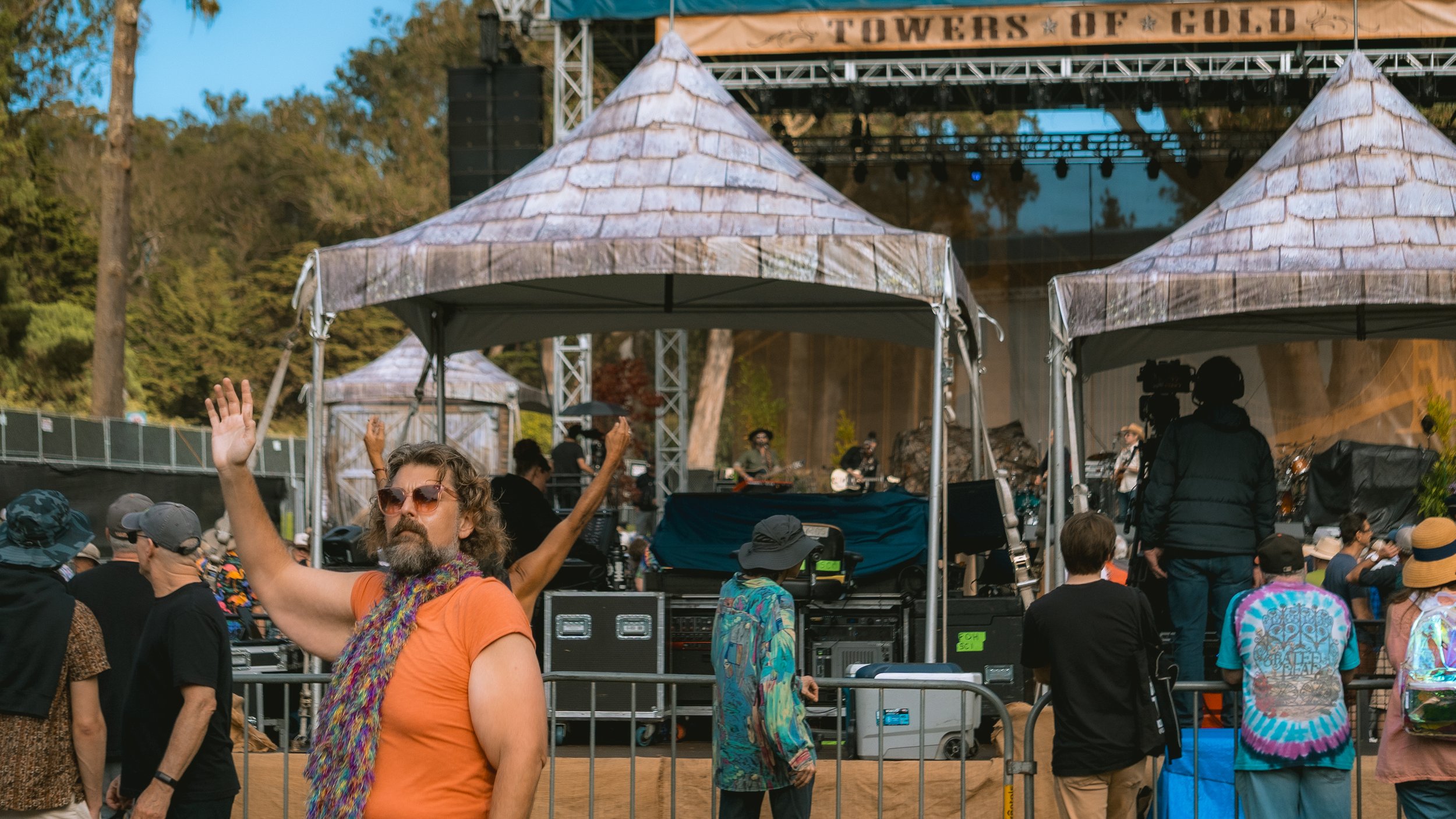 A crowd at an outdoor music festival with a stage and tents, with trees in the background. A man with sunglasses and an orange shirt is in the foreground, raising his hand. Other people are standing and watching the performance.