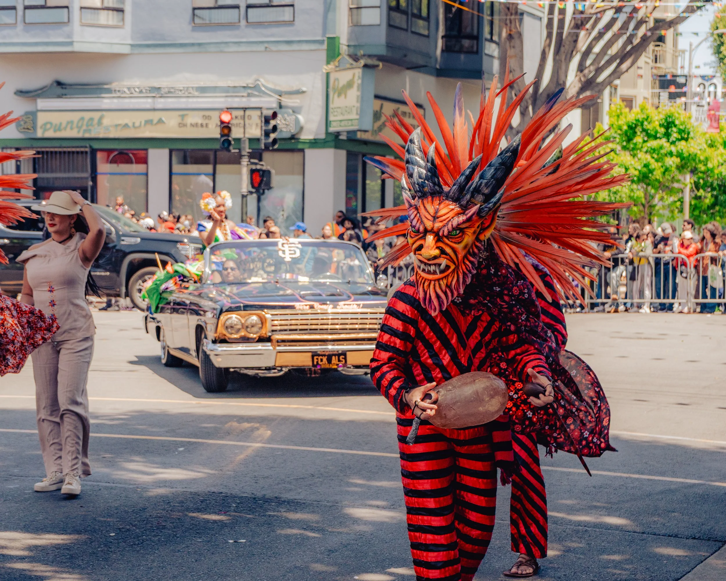 A person dressed in a detailed, colorful demon mask with large spikes and horns, wearing a red and black striped costume, participating in a parade on a city street. The background includes vintage cars, spectators, and parade performers.