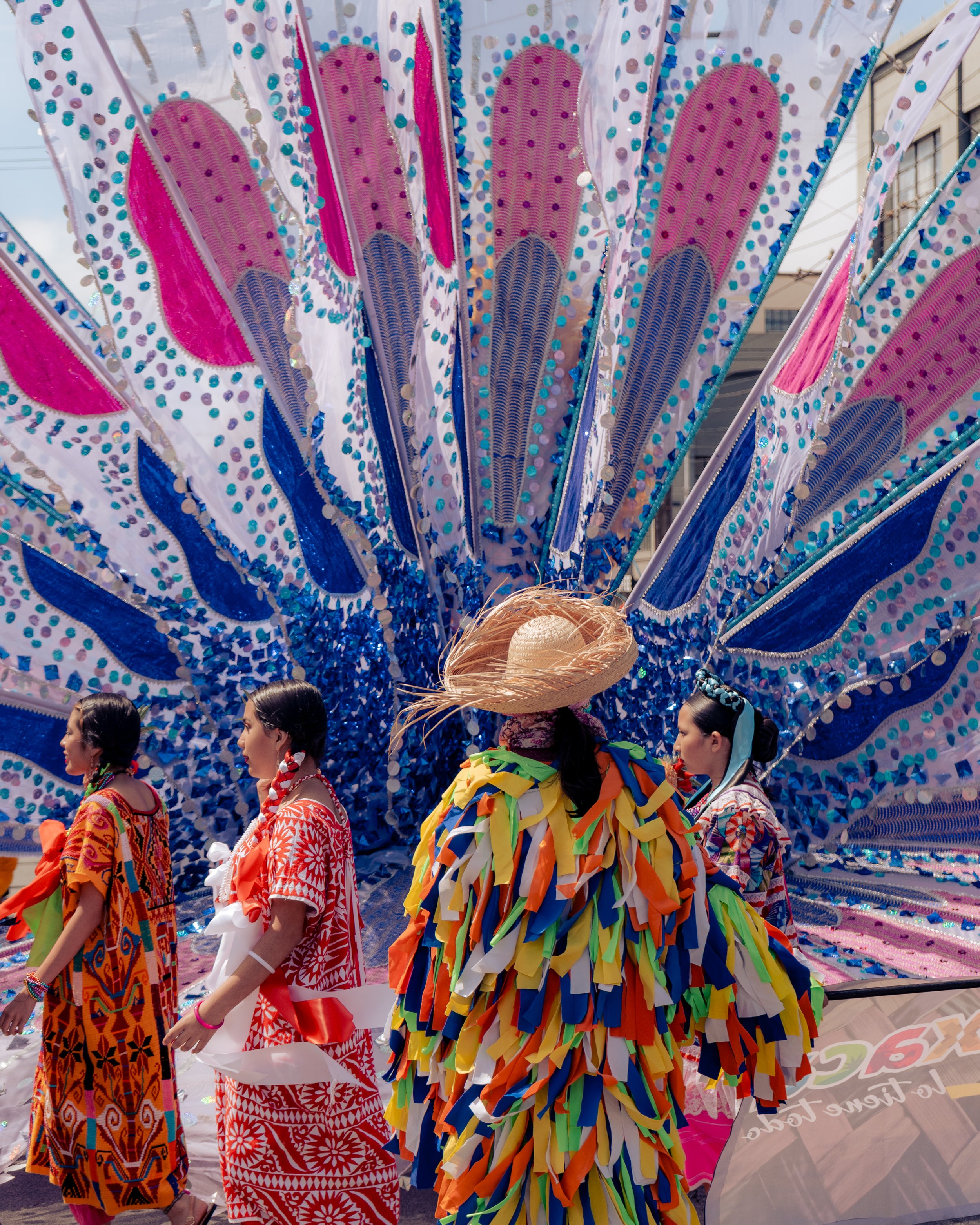 People in colorful traditional costumes marching in a parade with a decorated float in the background.