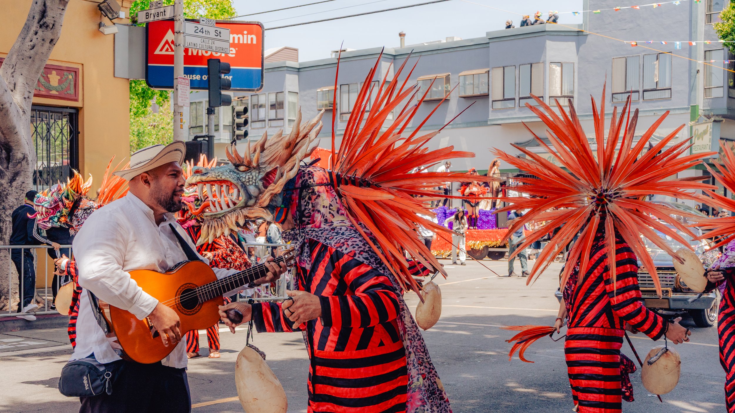 Man playing guitar in parade with performers wearing red and black striped costumes with large orange feathered headdresses and dragon masks.