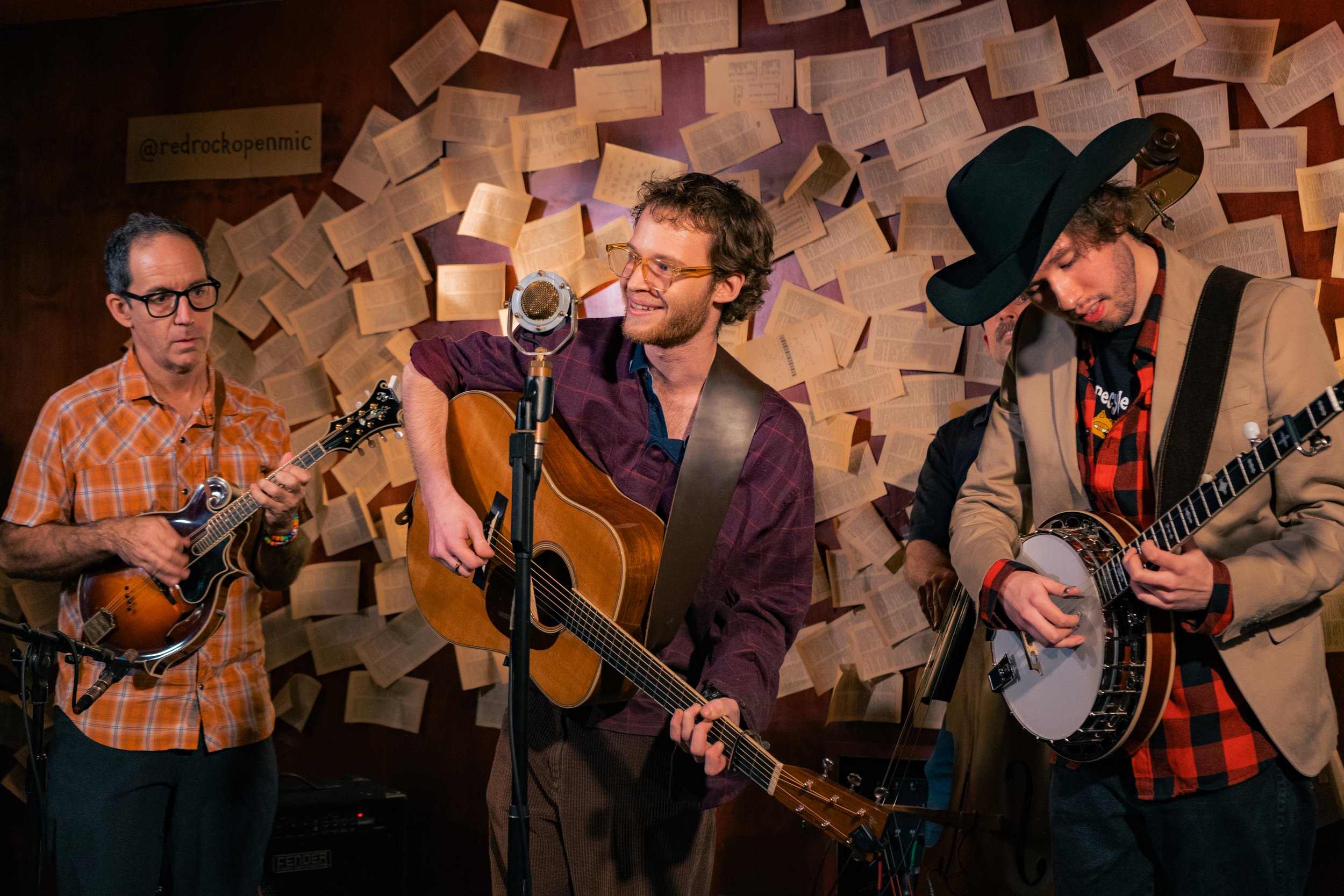 Three musicians playing stringed instruments in a room decorated with pages of printed text on the wall.