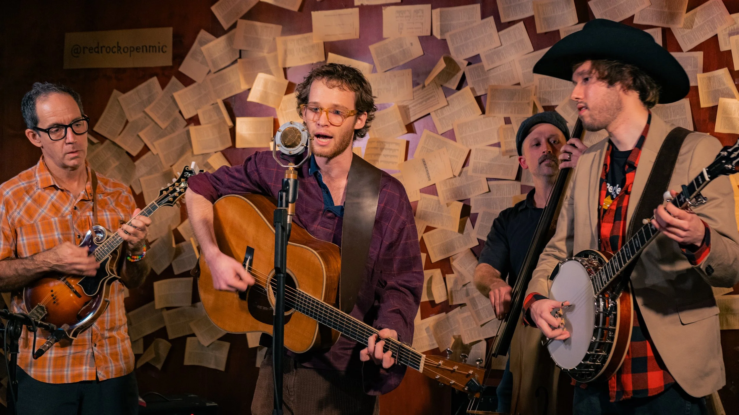 Four men playing guitars and singing on stage, with pages of sheet music or notes on the wall behind them.
