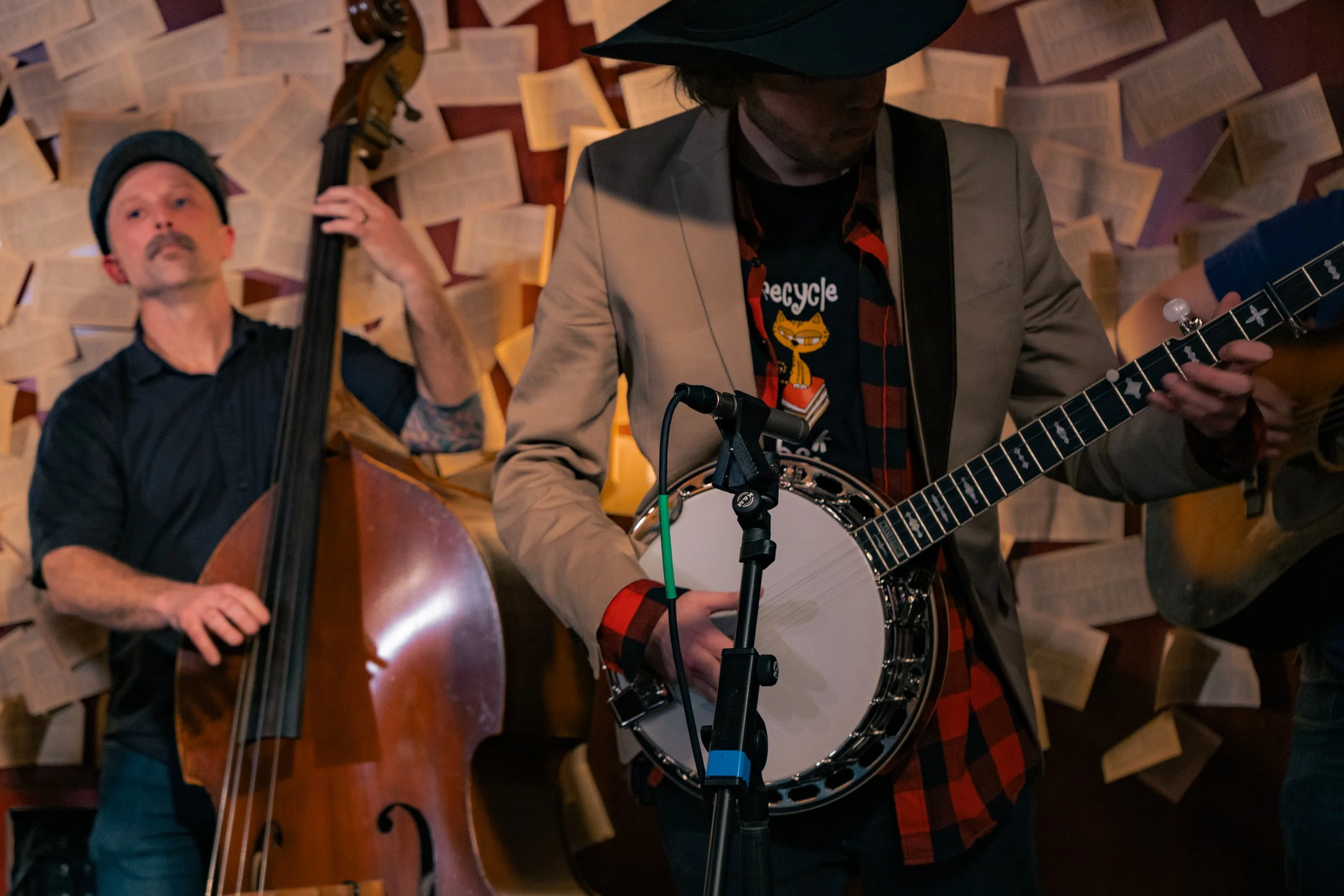 Two musicians performing indoors. One playing a double bass and the other playing a banjo, with pages of sheet music on the wall behind them.
