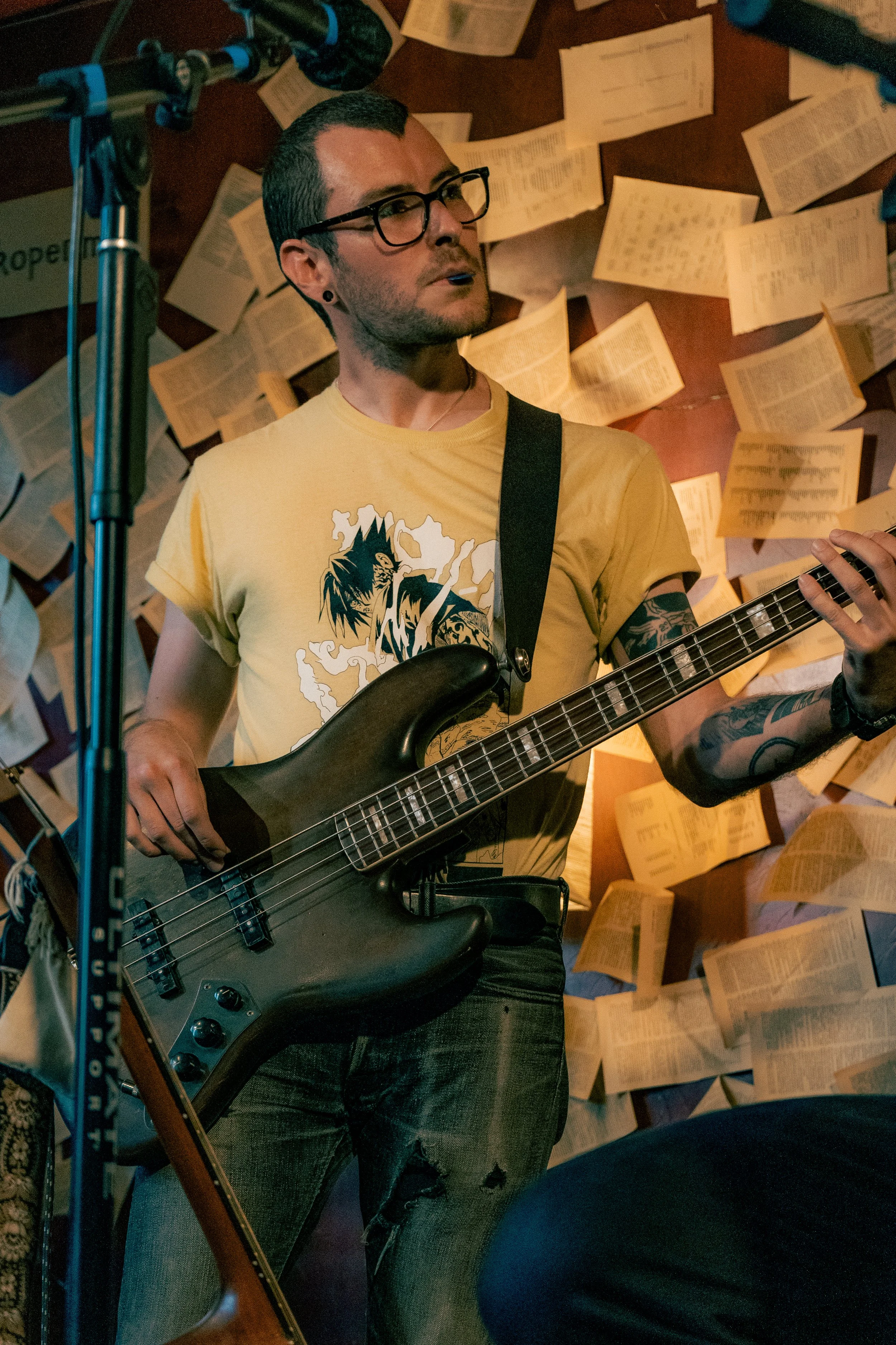 A man with glasses, tattoos, and a yellow t-shirt playing an electric bass guitar, in front of a wall covered with open books or papers, possibly during a performance or rehearsal.