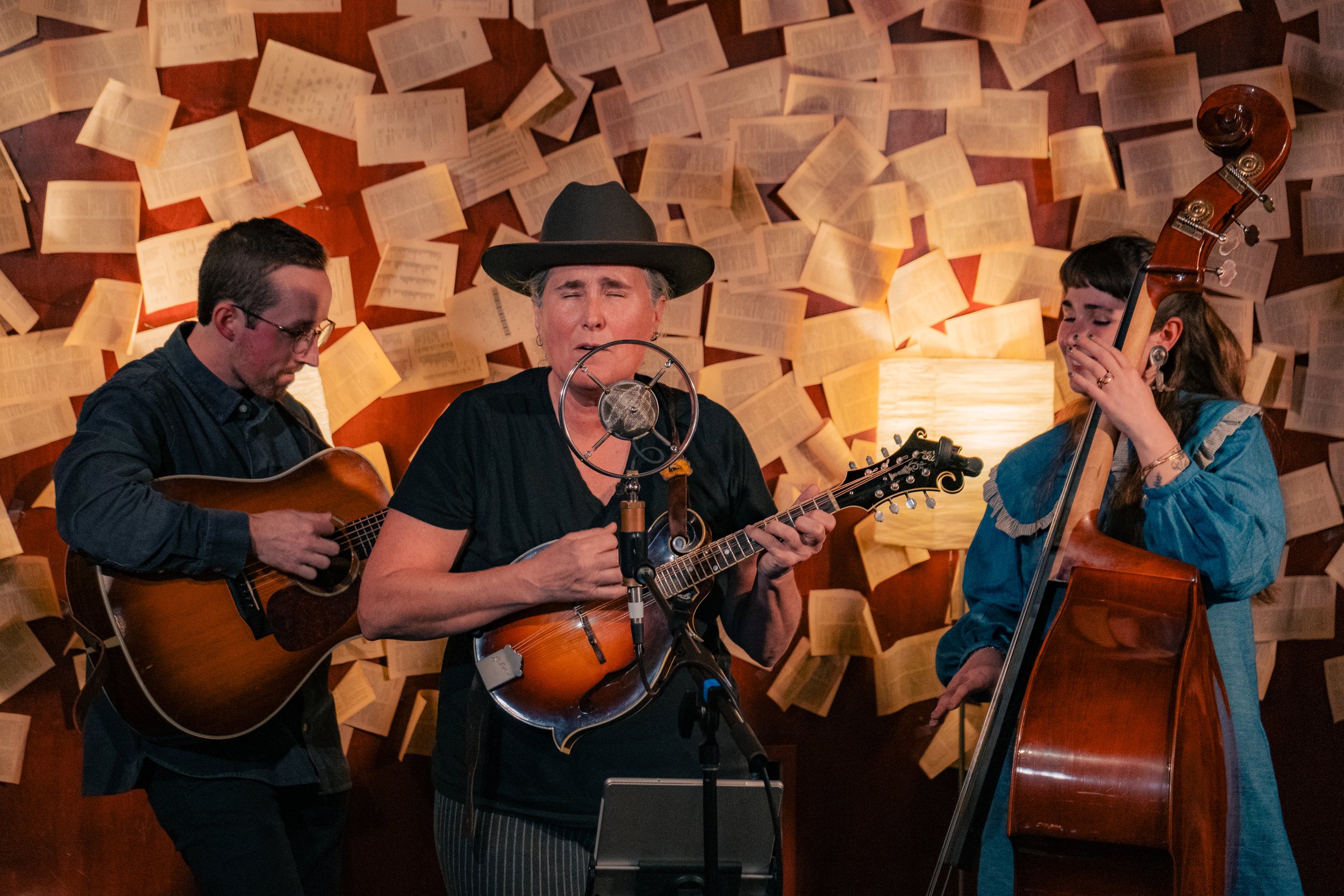 Three musicians performing in front of a wall covered with open books. The musician in the center sings into a vintage microphone while playing a small string instrument, flanked by a guitarist on the left and a double bass player on the right.
