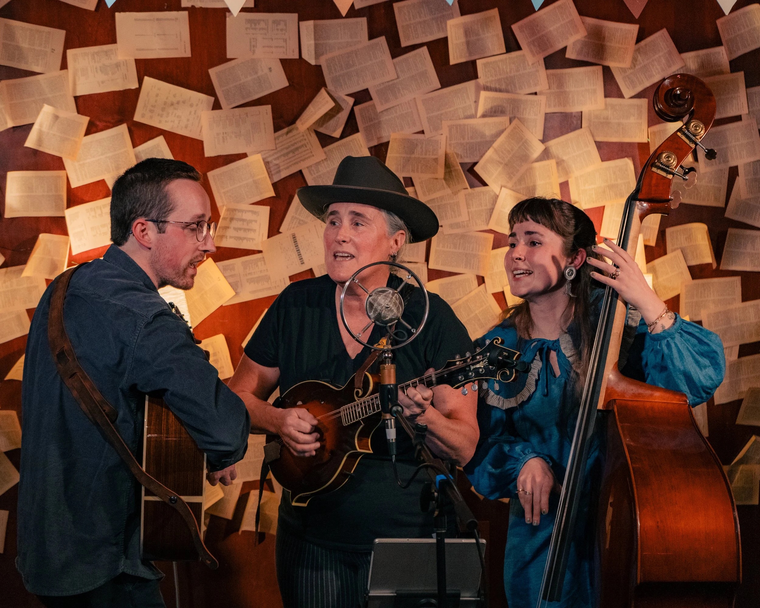 Three women singing and playing guitars and a bass in front of a backdrop covered with open books.