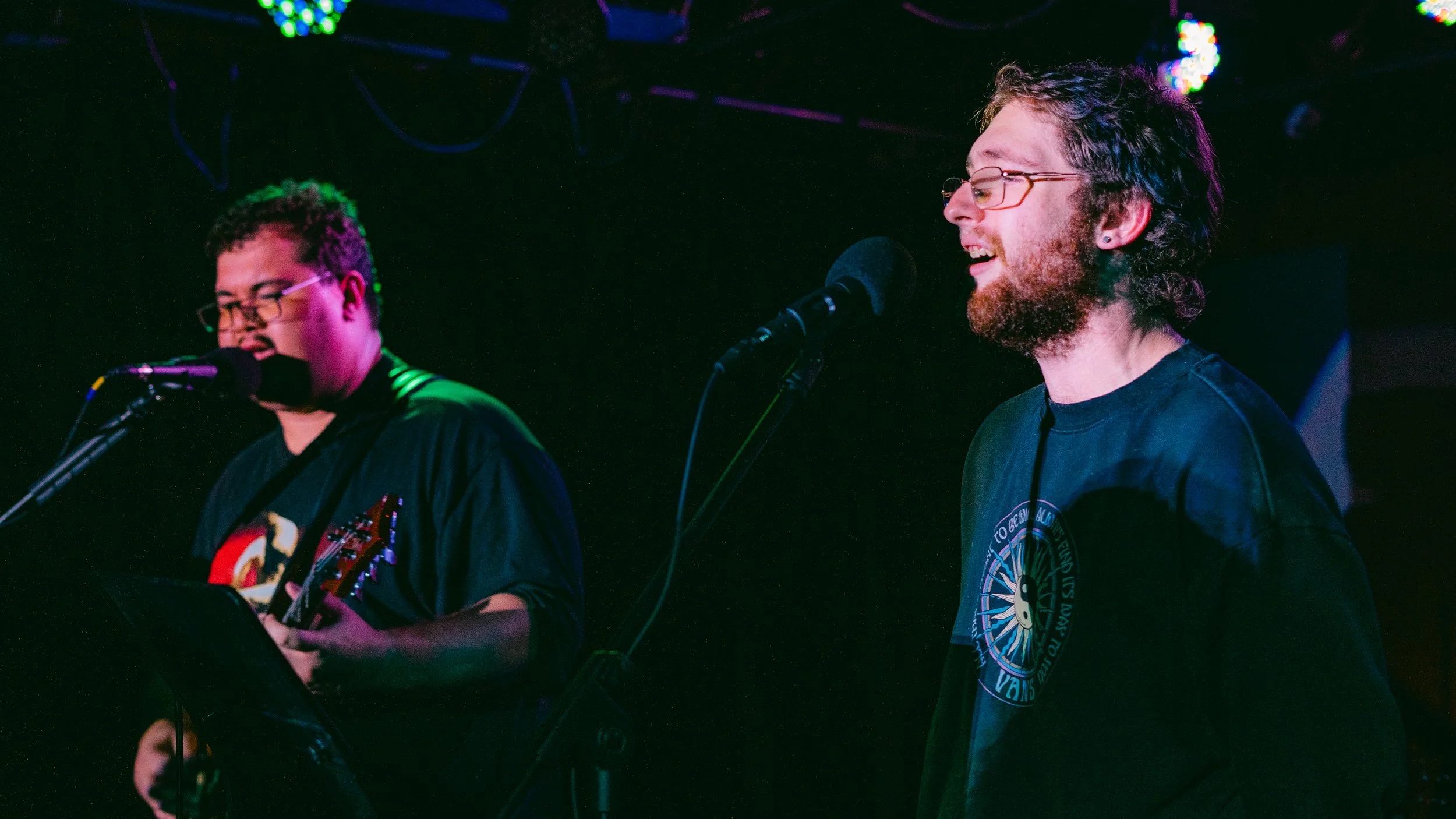 Two young men performing on stage, one singing into a microphone and playing a guitar, and the other singing into a microphone with closed eyes. The stage is lit with colorful lights.