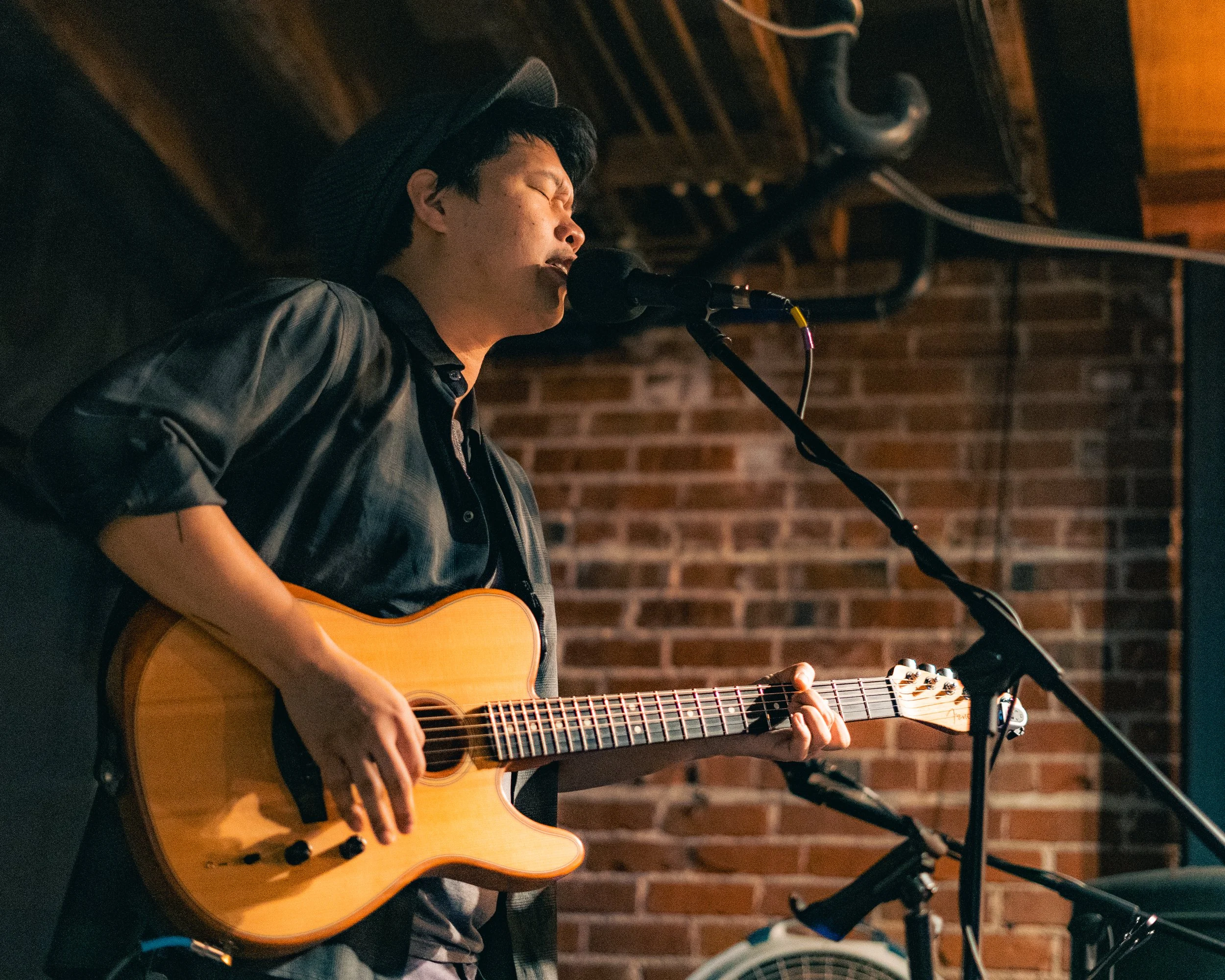 A young man singing into a microphone while playing an acoustic guitar on stage with a brick wall background.