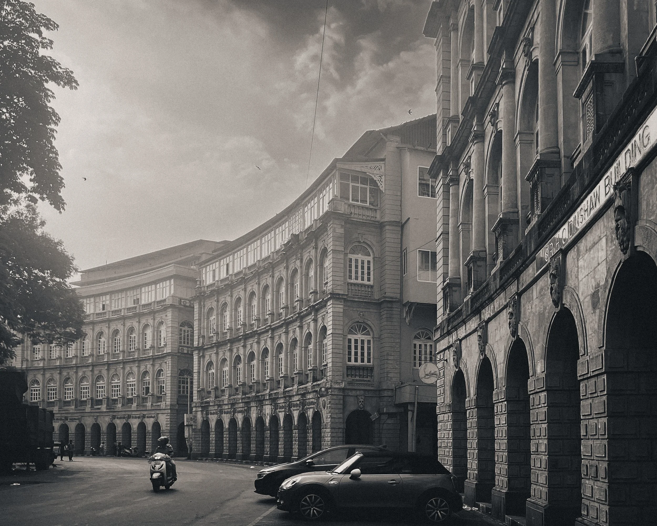 Black and white photograph of an old city street with ornate multi-story buildings, parked cars, and a motorcycle. The sky is cloudy, and there are a few birds flying.