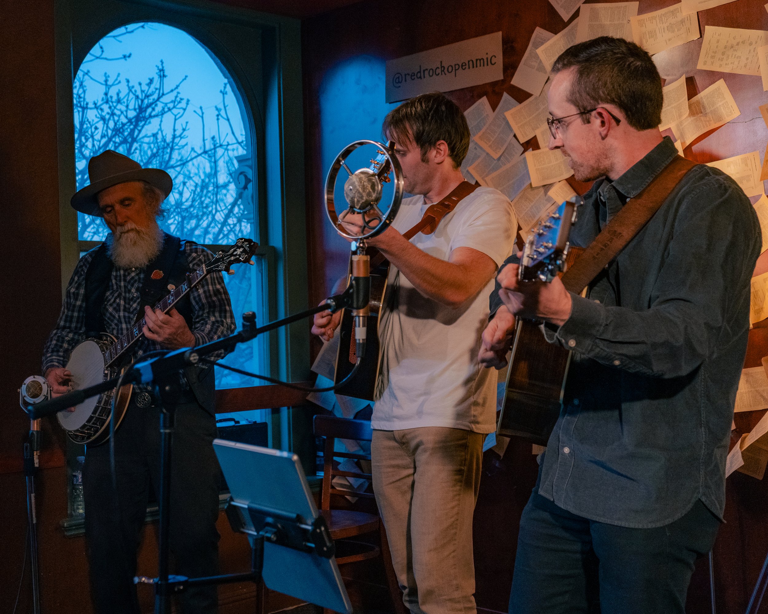 Three men playing musical instruments indoors, with a window showing tree branches outside. One man is playing a banjo, another is playing an acoustic guitar, and the third man appears to be adjusting a microphone.