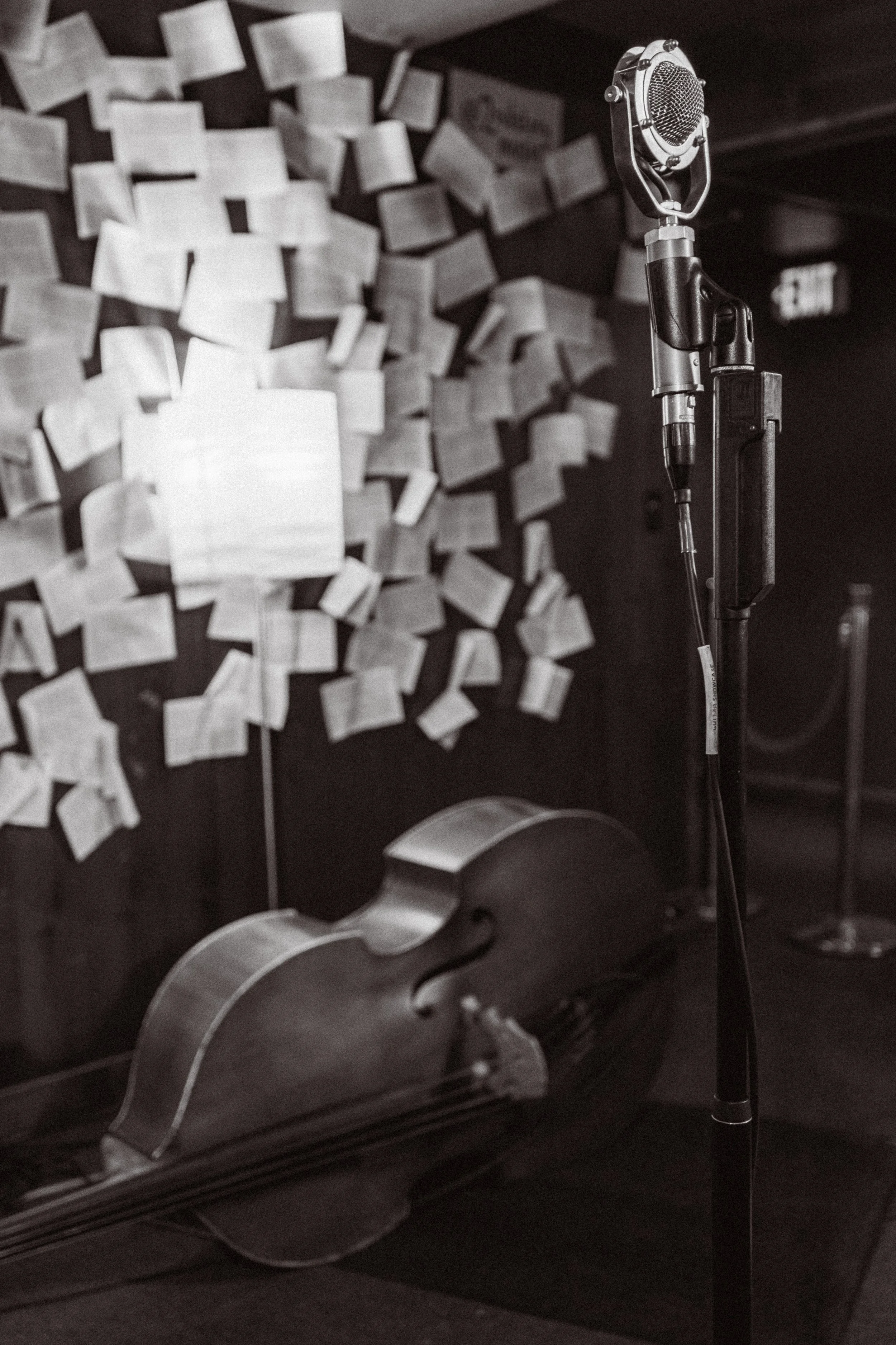 A microphone stand with a vintage microphone, a cello lying on the floor, and a wall covered with numerous notes or pieces of paper in the background.