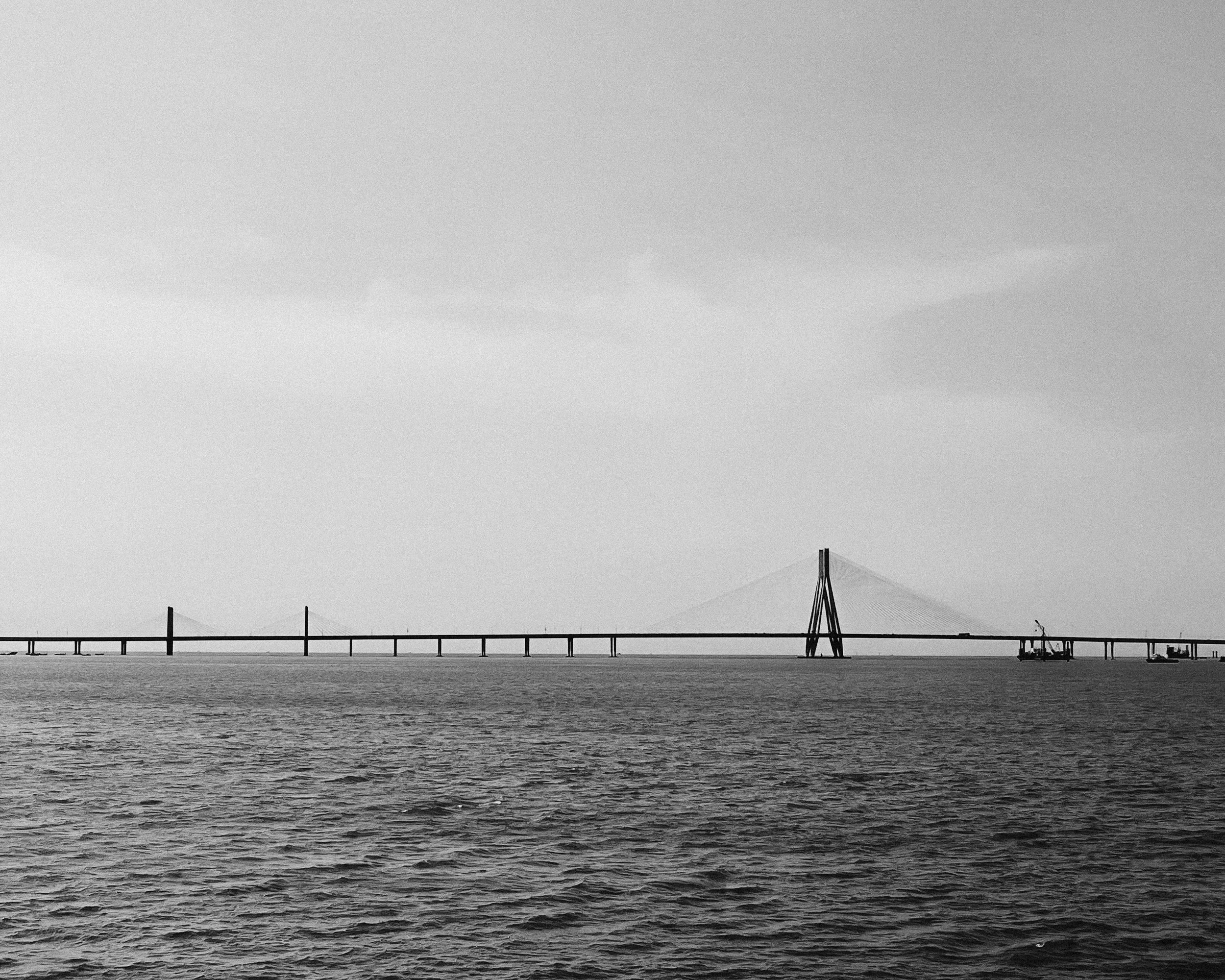 Black and white photo of a long bridge over water with a cable-stayed section and a distant bridge in the background.