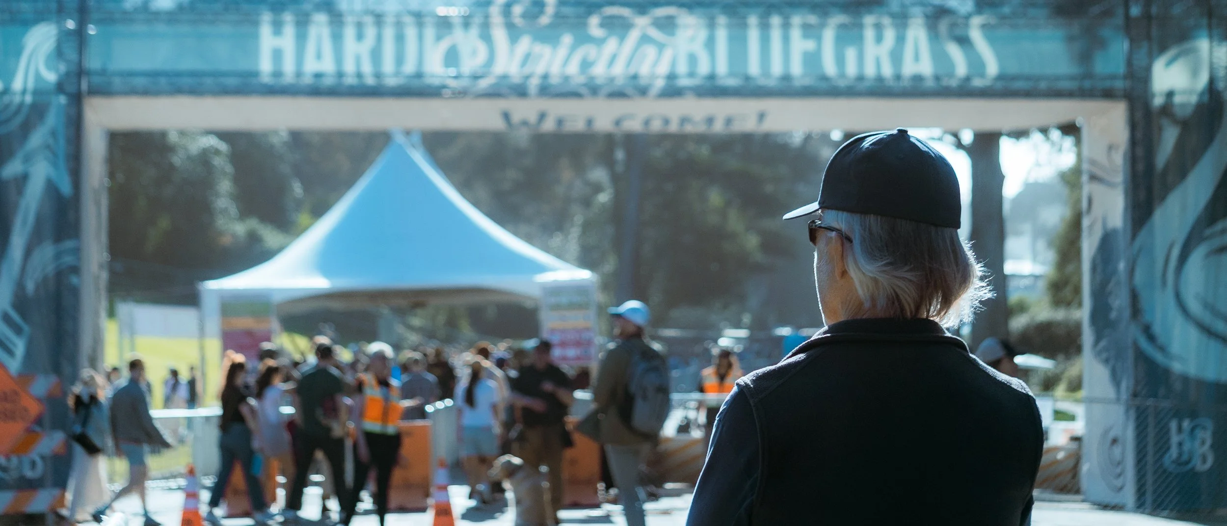 A person with gray hair, wearing a black cap and glasses, seen from behind, stands at the entrance of a public event or festival with people walking and gathered near a tent and barricades.