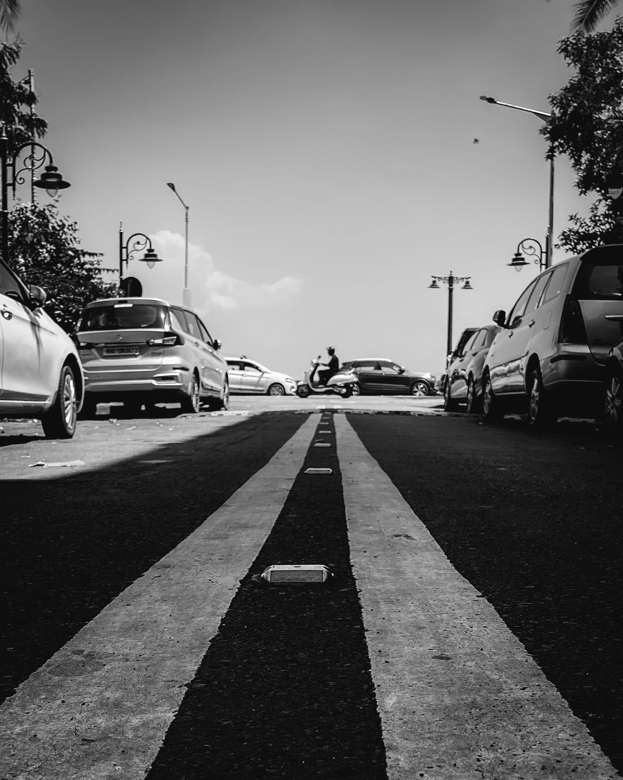 Black and white photo of a street with parked cars on both sides and a motorcyclist in the distance, taken from a low angle focusing on the road markings.