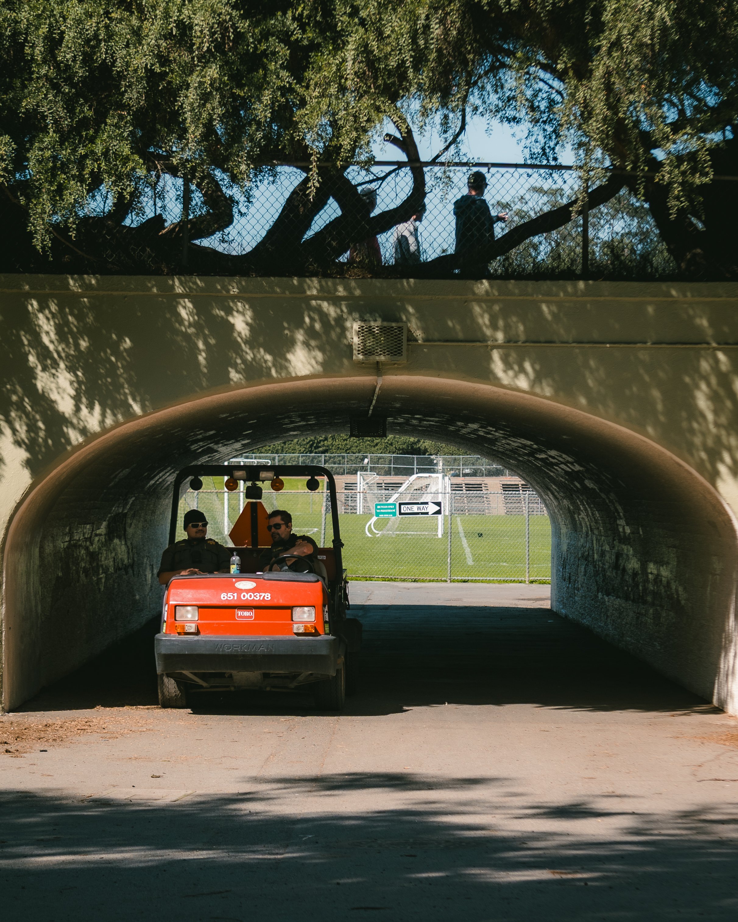 Two police officers in uniform riding in a small orange utility vehicle under a bridge, with a chain-link fence and trees above.