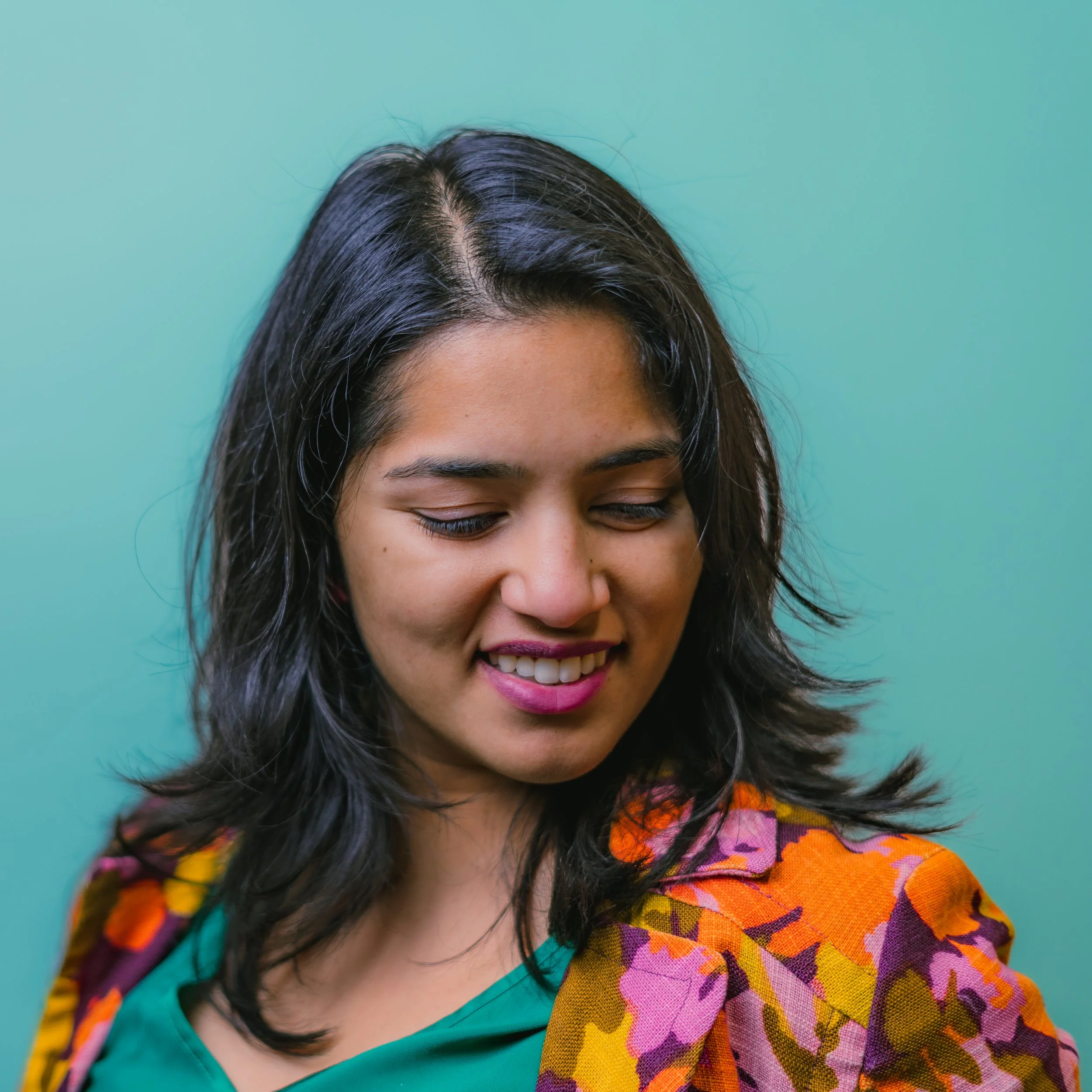 A young woman with dark hair smiling and looking down, wearing a colorful patterned jacket over a teal top, against a teal background.